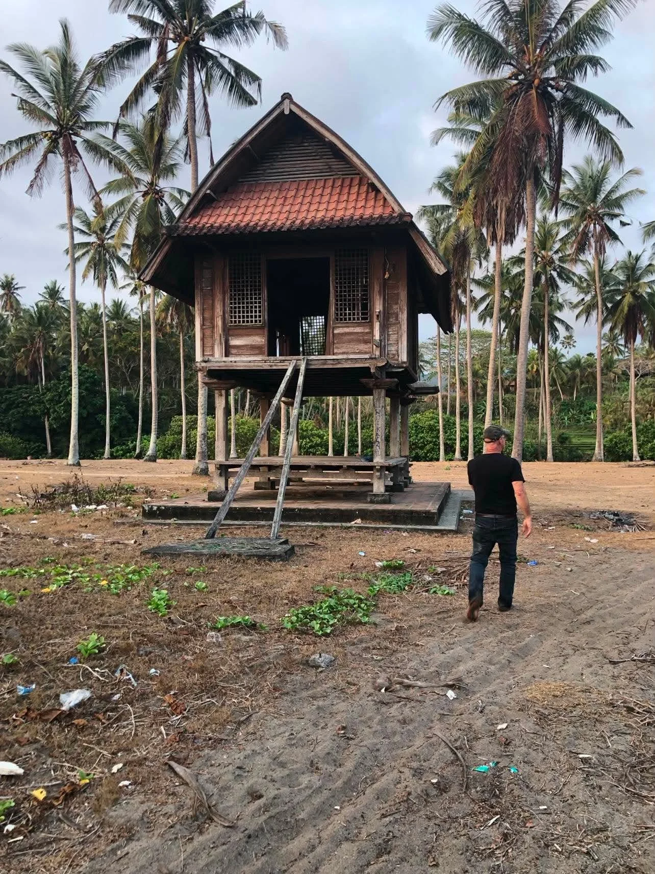 With his back to the camera, Al walks across a sandy path toward a home on stilts in Indonesia.
