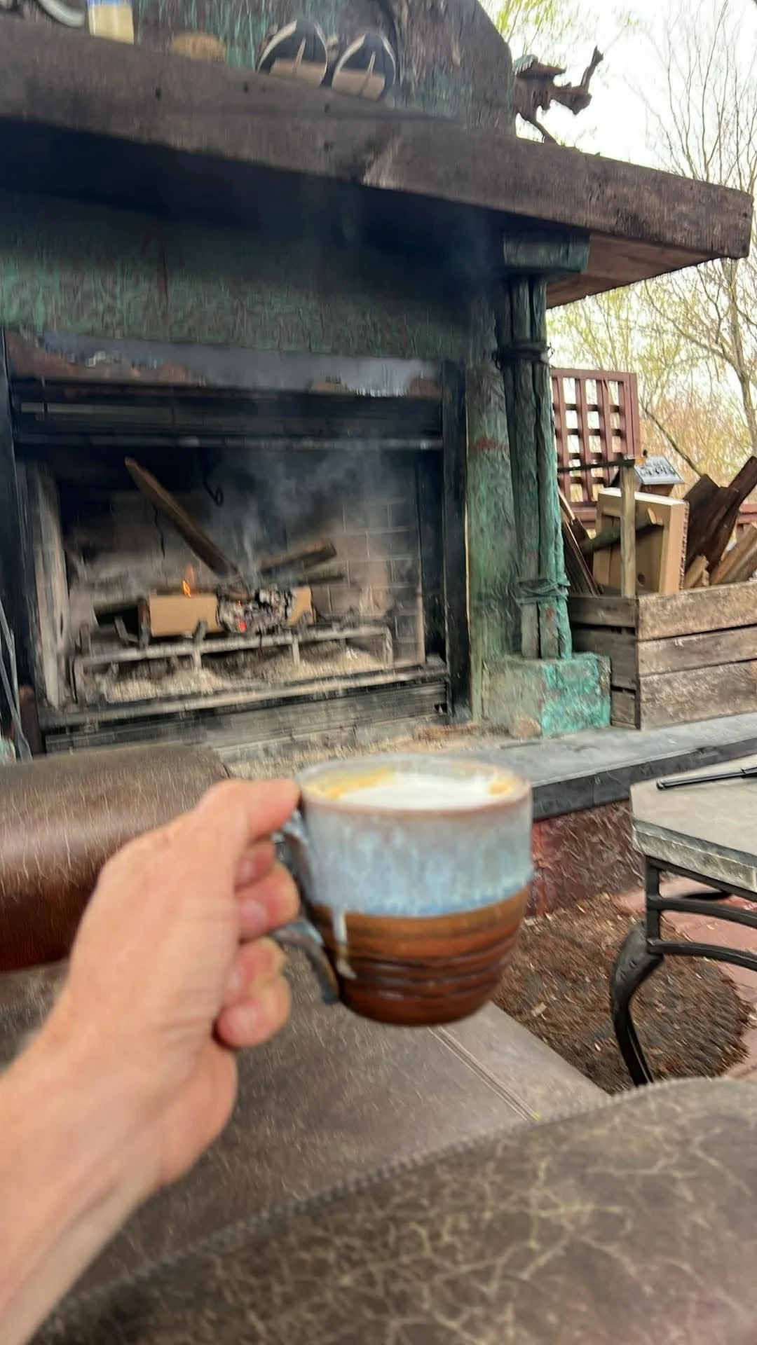An outstretched hand holds an elegant ceramic coffee cup brimming with a hot latte. In the background, a fire burns down in a large outdoor fireplace.