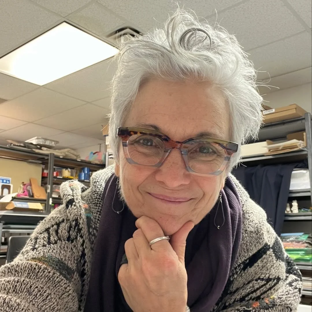 Deb, with short gray hair, stylish glasses, and a purple scarf, sits in front of her art studio and smiles into the camera with her hand on her chin.