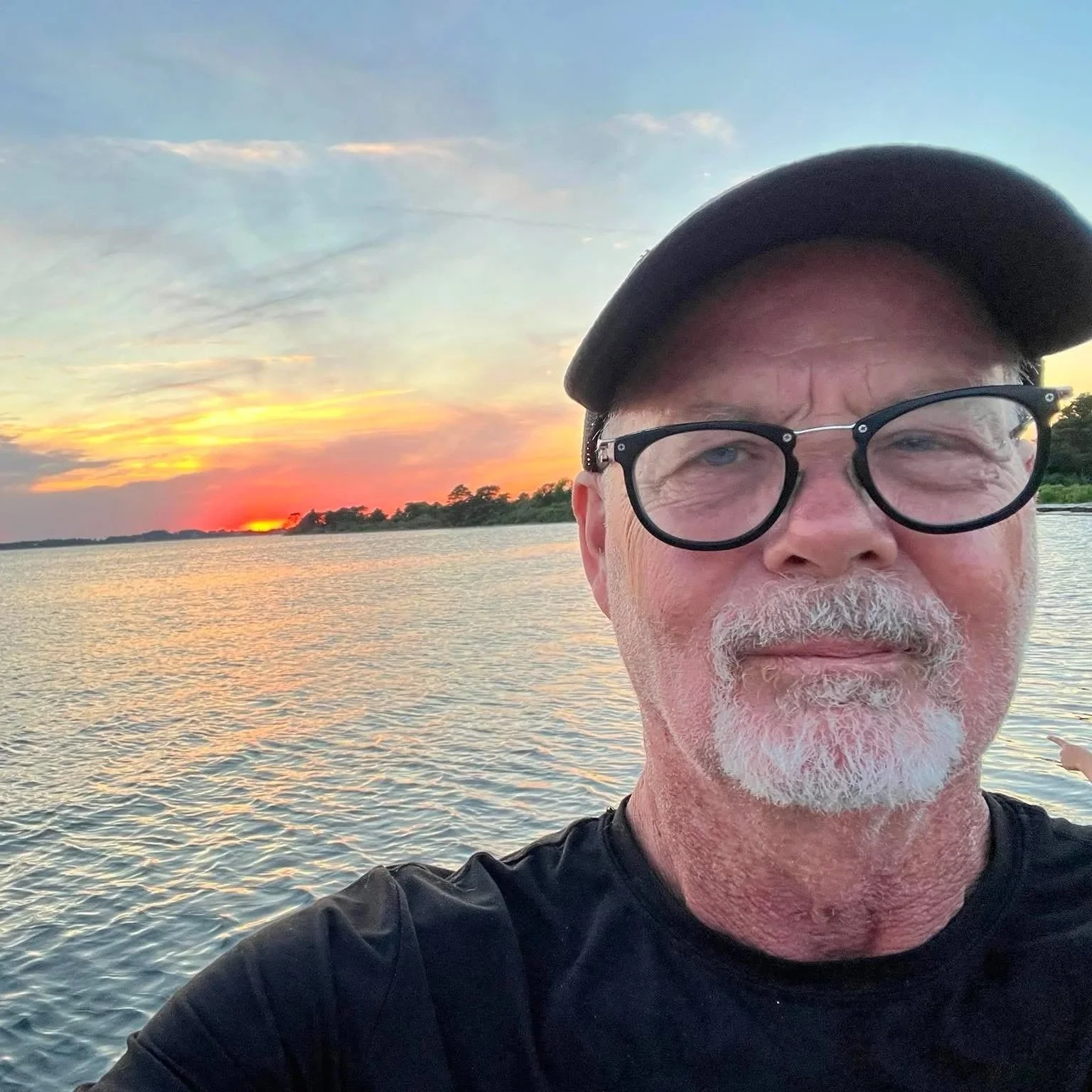 Al stands in front of a sunset over a body of water, wearing a black baseball cap, black-rimmed glasses, and a black shirt.