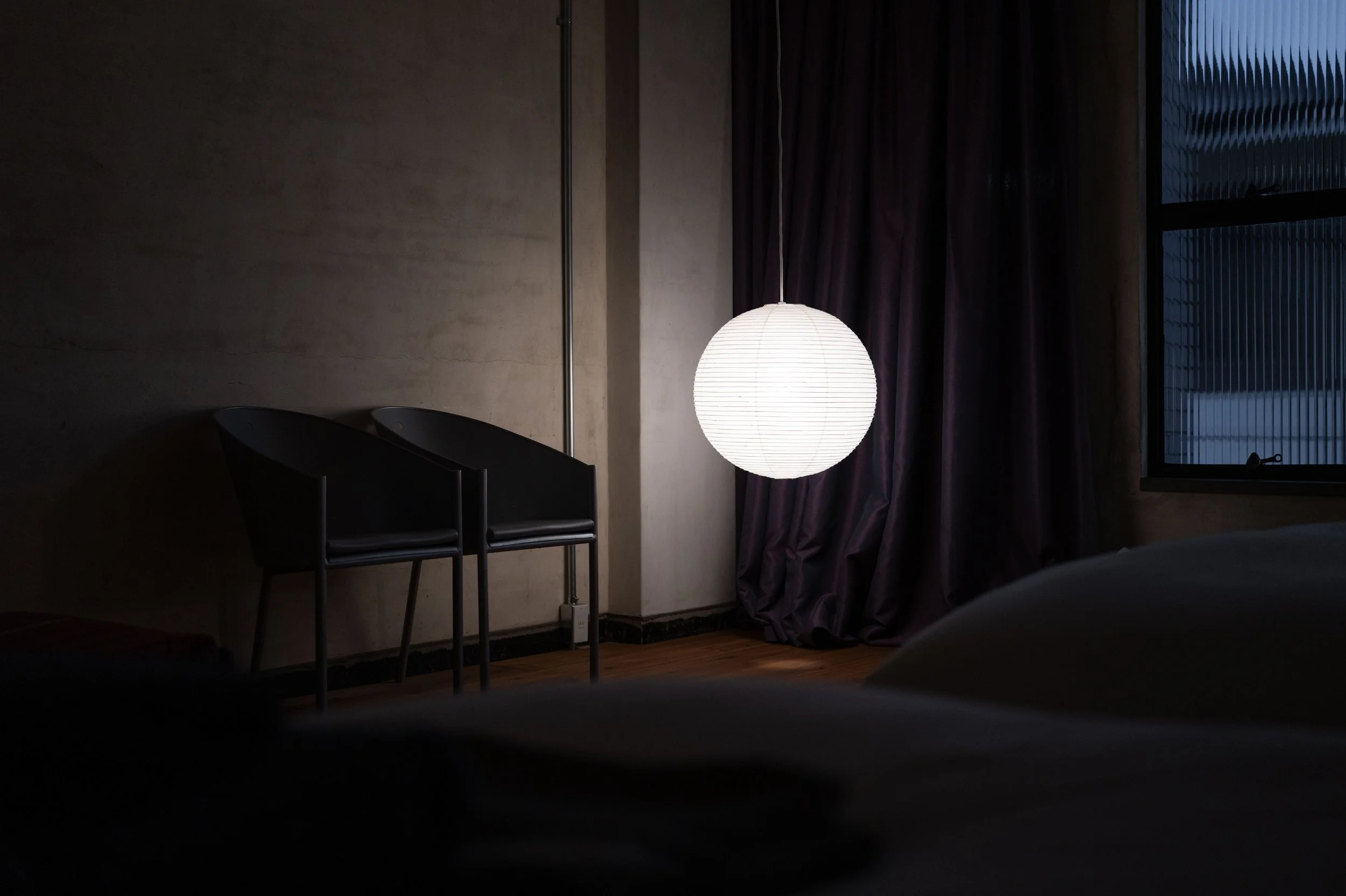 A dimly lit room with a glowing paper lantern hanging from the ceiling, dark chairs tucked against the wall, a window with curtains, and a partial view of a bed or furniture in the lower right corner.