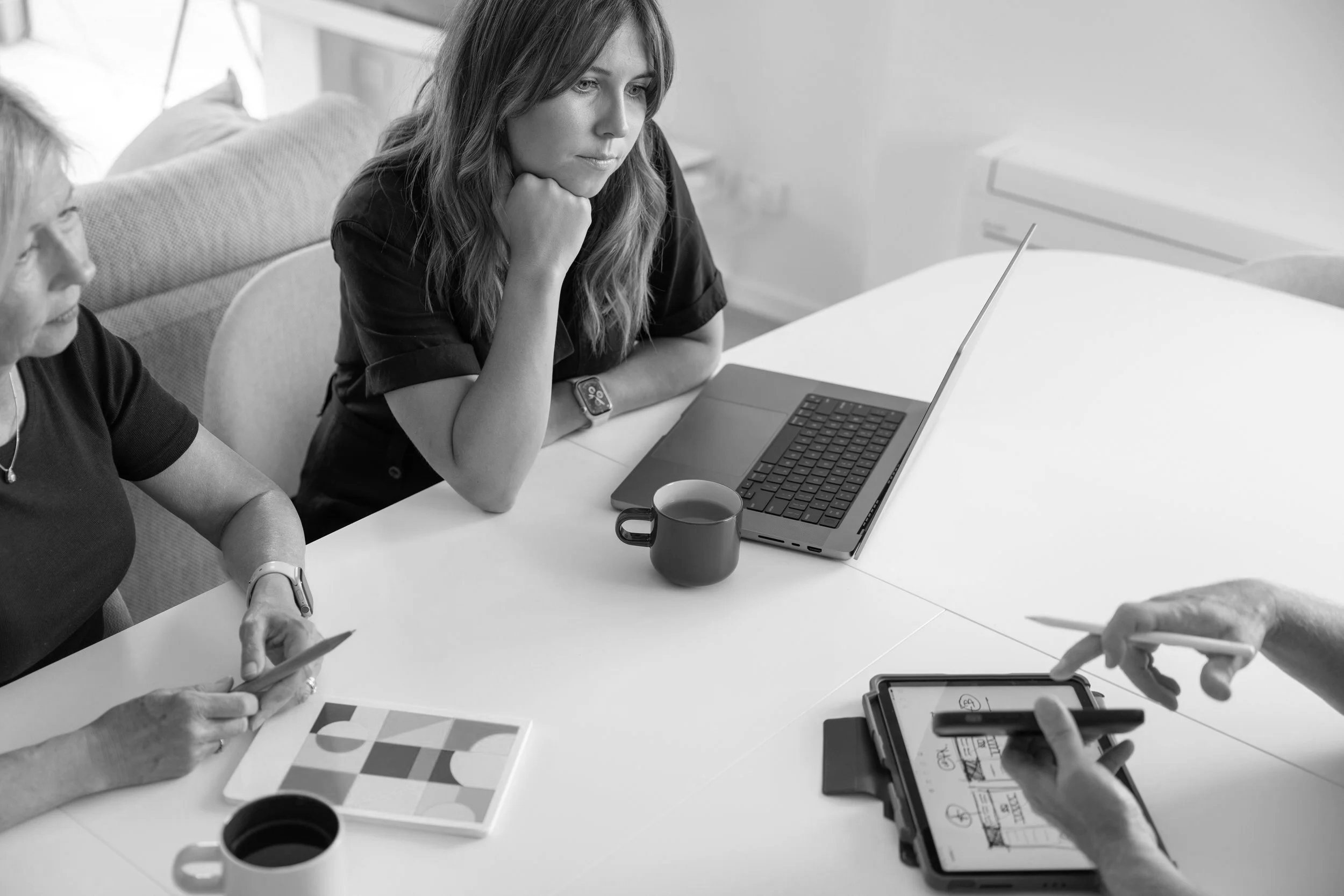 Two women are sitting at a table working on electronic devices such as smartphones and laptops, with papers and coffee mugs present, in a modern indoor setting.