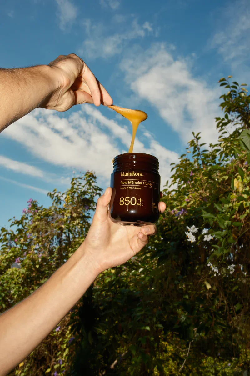 A person holding a jar of Manukora raw Manuka honey with a spoonful dripping into the jar, outdoors during daytime with a blue sky and green foliage in the background.