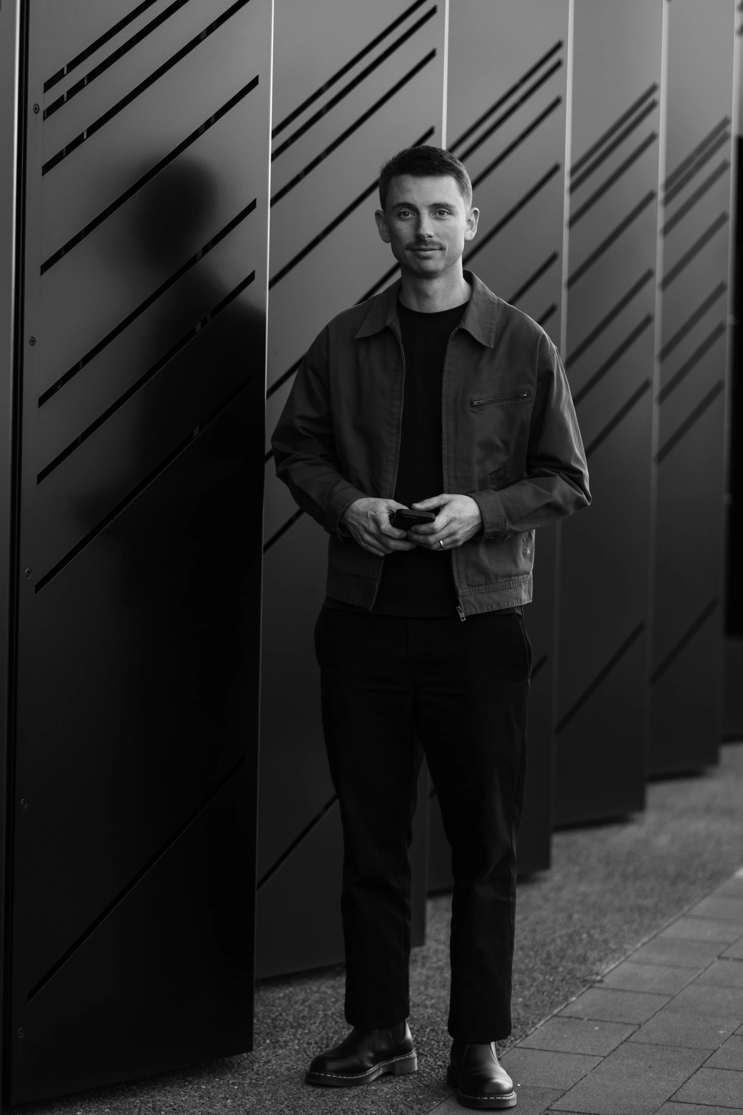 Andy standing outdoors by modern lockers, wearing a jacket and holding a phone, in black and white.