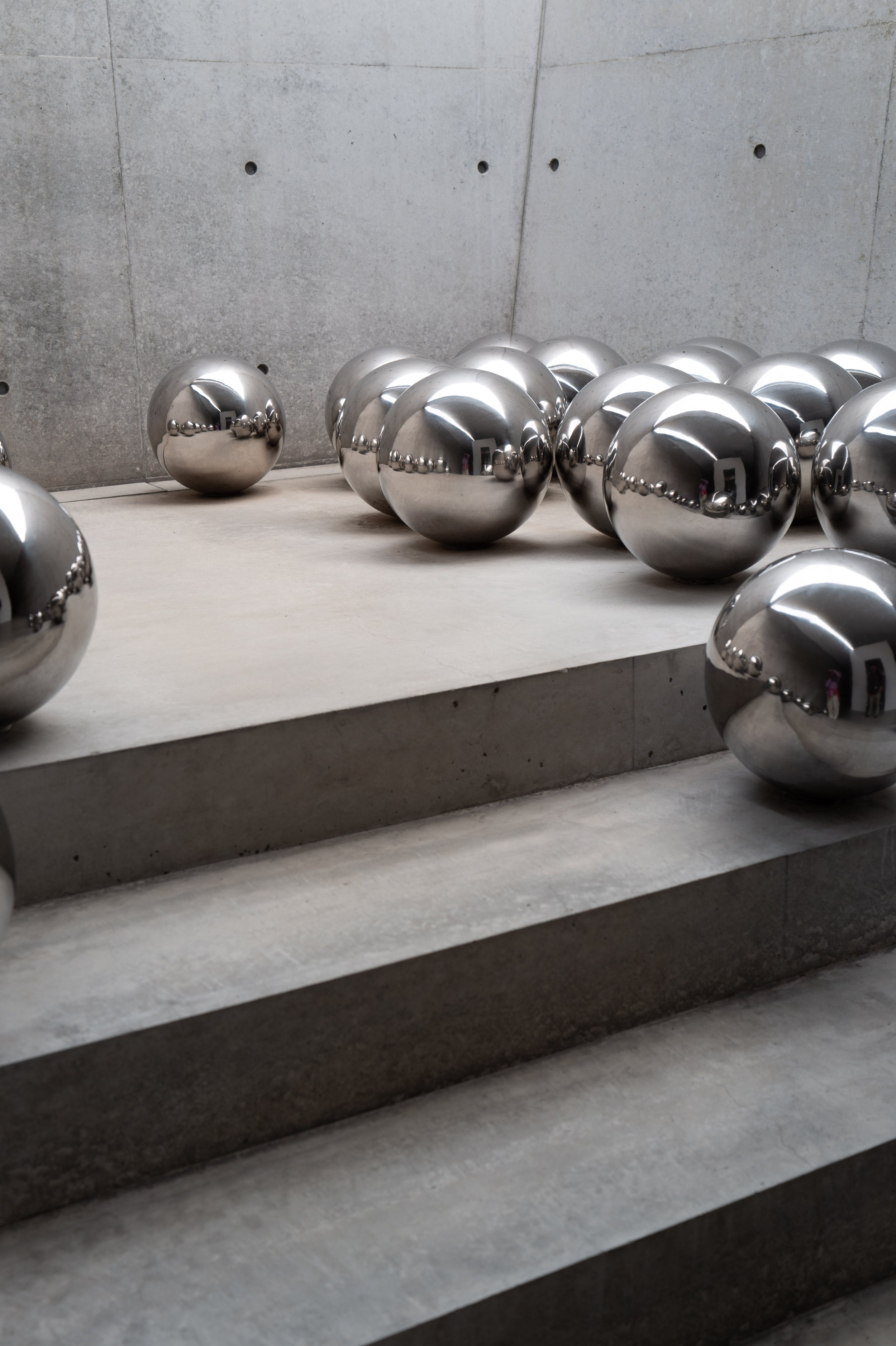 Several shiny metallic spheres arranged on a tiered concrete surface with a gray concrete wall in the background.