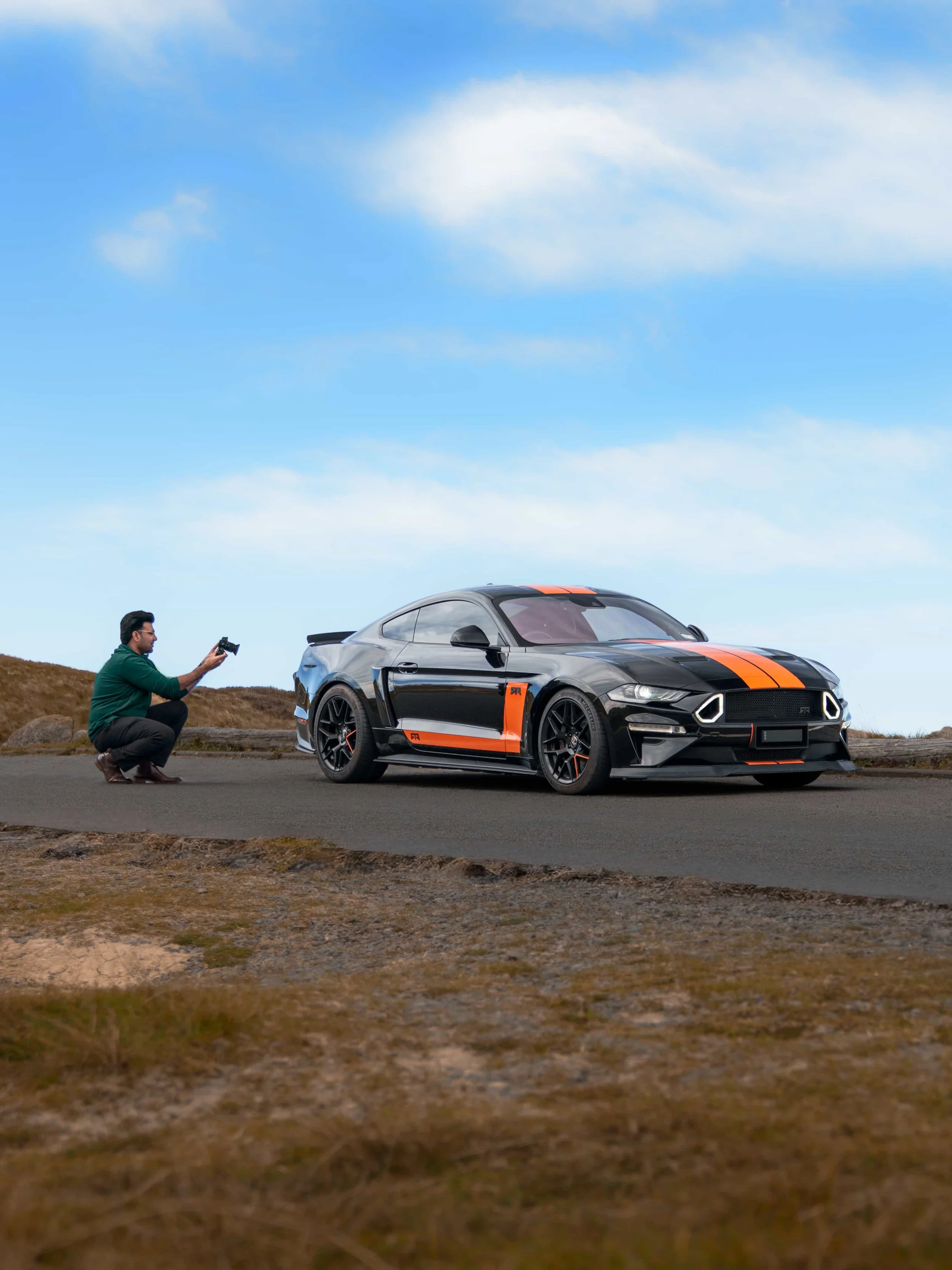 A man kneeling on the ground taking a photo of a black sports car with orange racing stripes on a road in a natural outdoor setting with blue sky and clouds.
