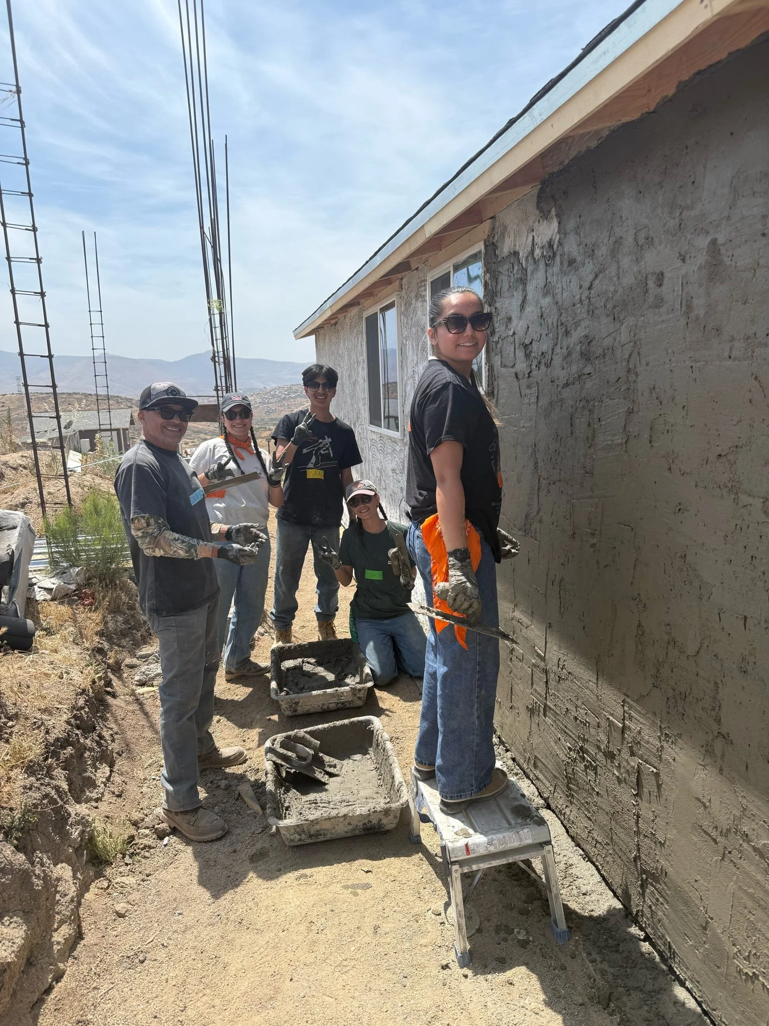Group of people working on a house construction or renovation project outdoors, with a partially finished exterior wall, tools, and construction materials around them, with mountains and a partly cloudy sky in the background.