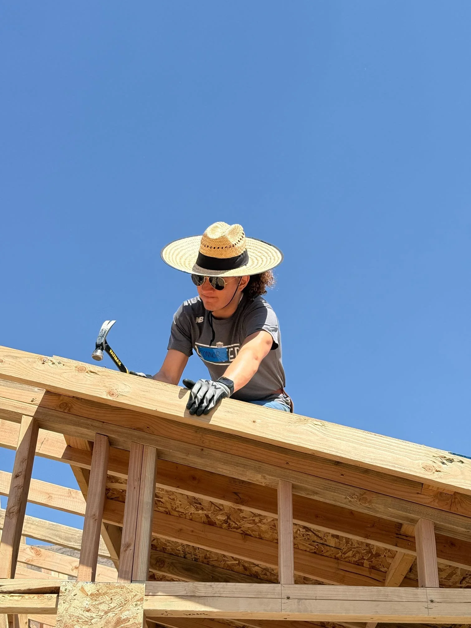 A construction worker wearing a wide-brimmed straw hat, sunglasses, a gray shirt, and gloves is working on a wooden structure against a clear blue sky.