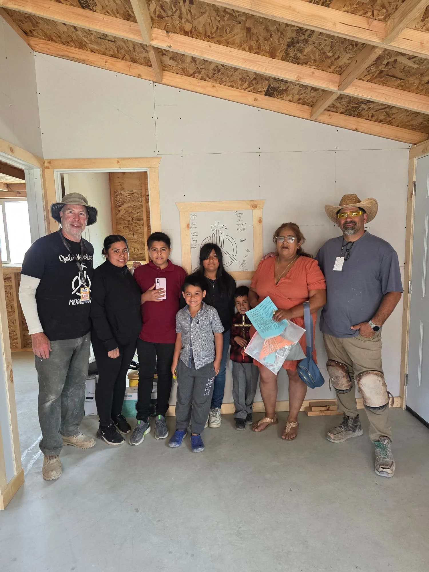 Family who is receiving the house standing inside great room, with visible wooden framing and drywall on one wall, and a sign with a cross and names on the wall behind them.