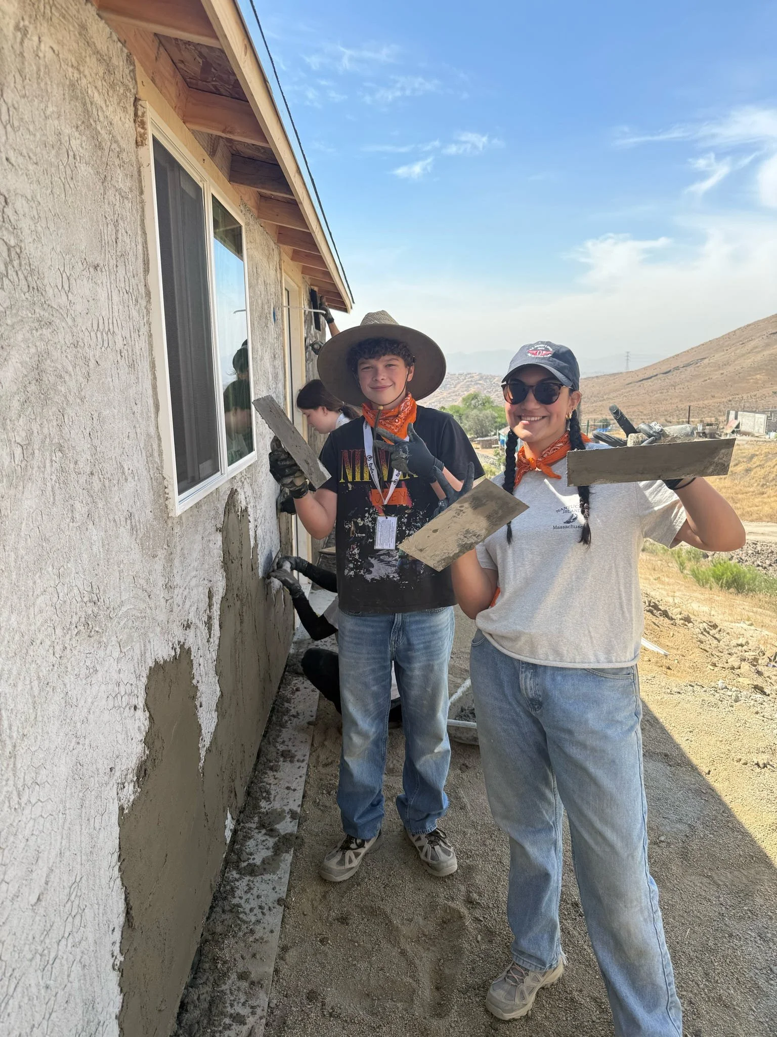 Two women working on a house exterior, applying stucco to a wall, with tools in their hands and wearing casual clothes, outdoors with a hilly landscape in the background.