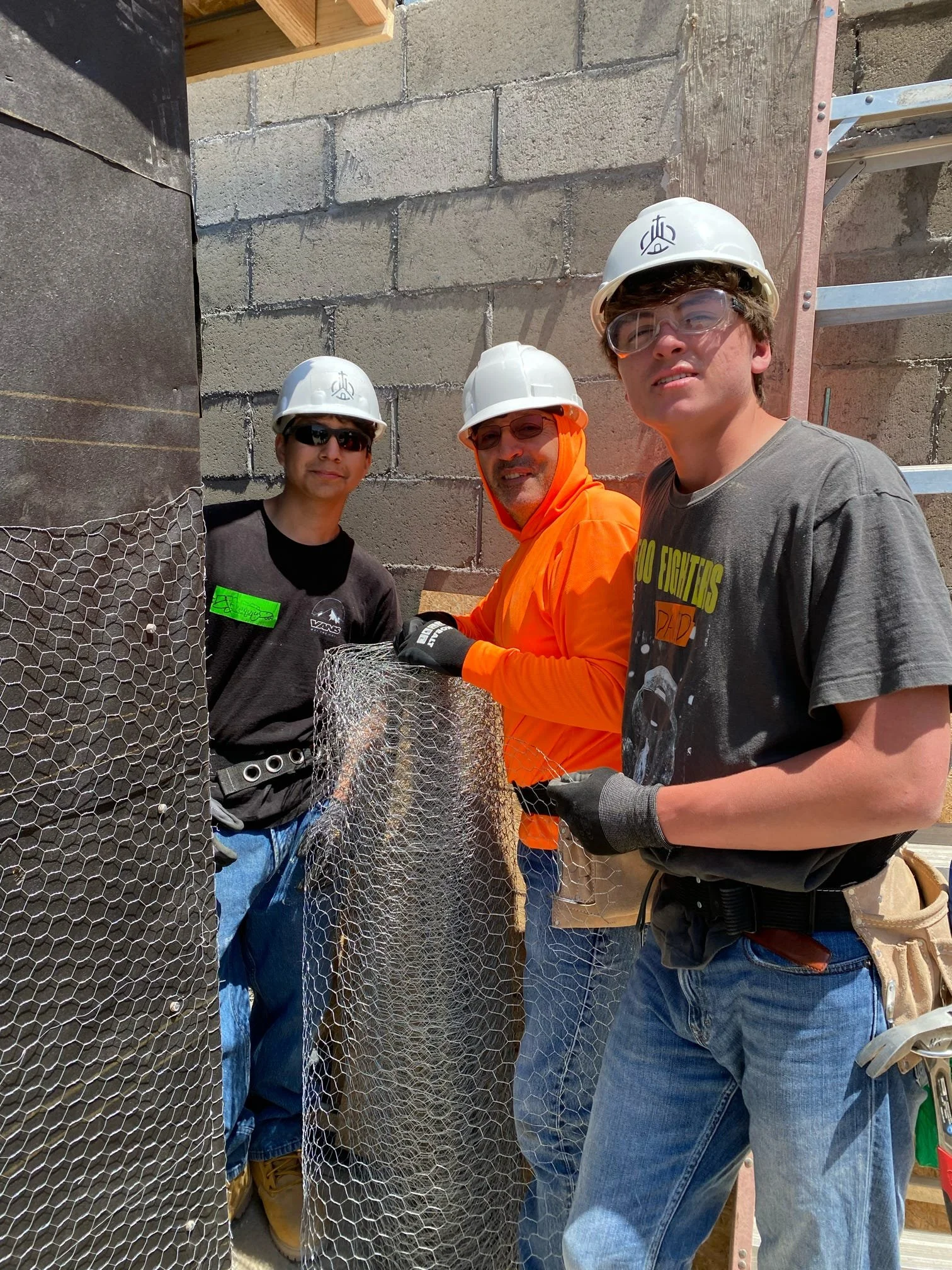 Three people wearing hard hats and safety glasses working on a construction site with cinder block walls and a ladder in the background, holding a roll of wire mesh.