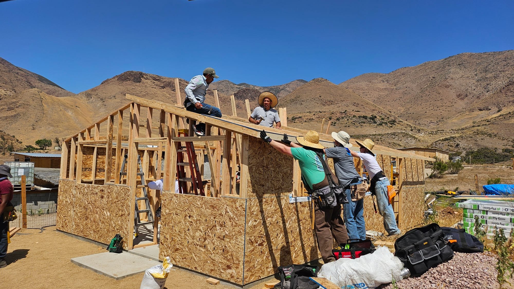 Construction workers building a house frame in a desert area with mountains in the background.