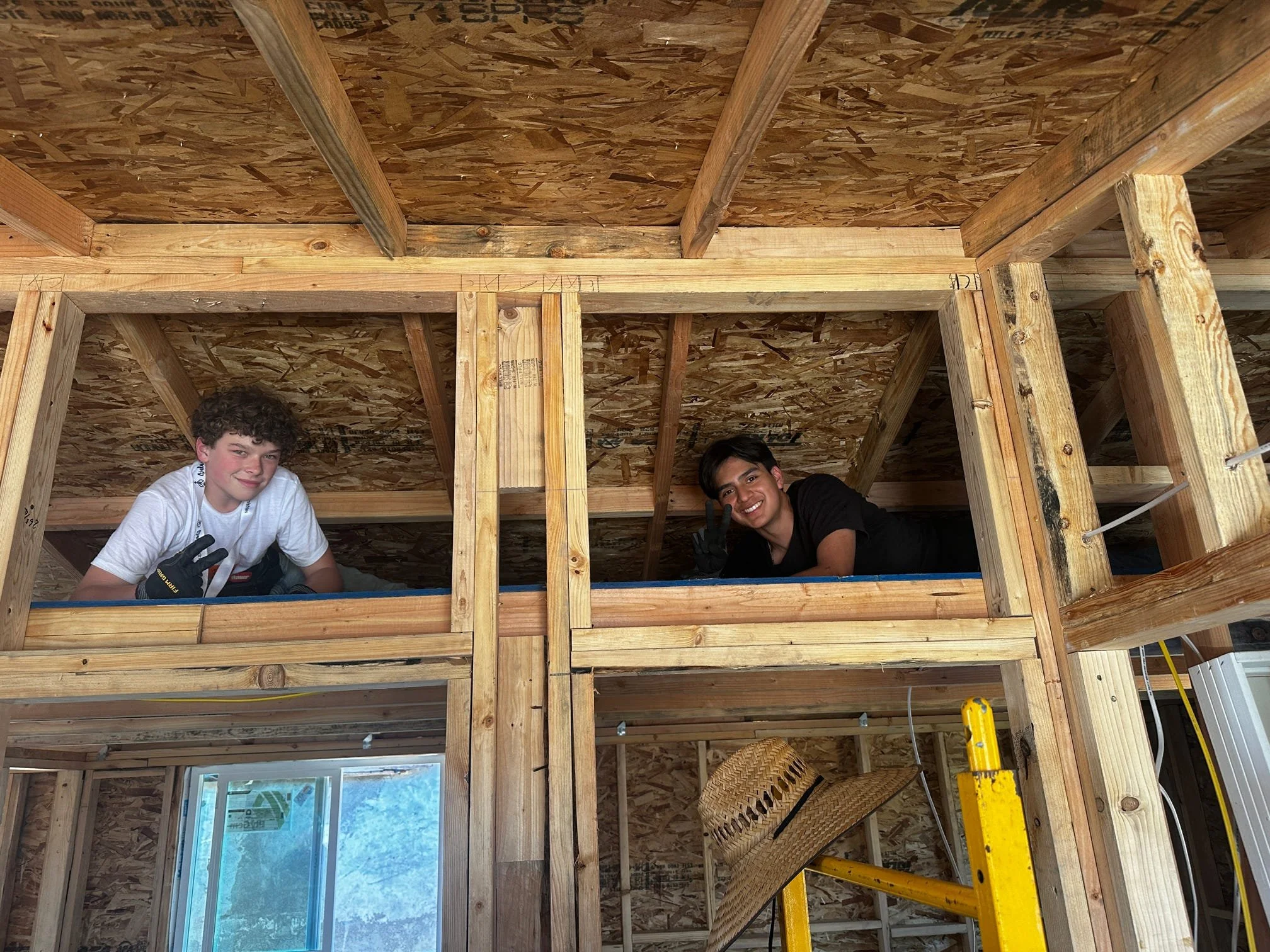 Two young men working on framing a room inside a house under construction. The room has wooden studs and OSB walls and ceiling. One man is wearing a white shirt and black gloves, and the other is wearing a black shirt. There are construction tools and materials around them.