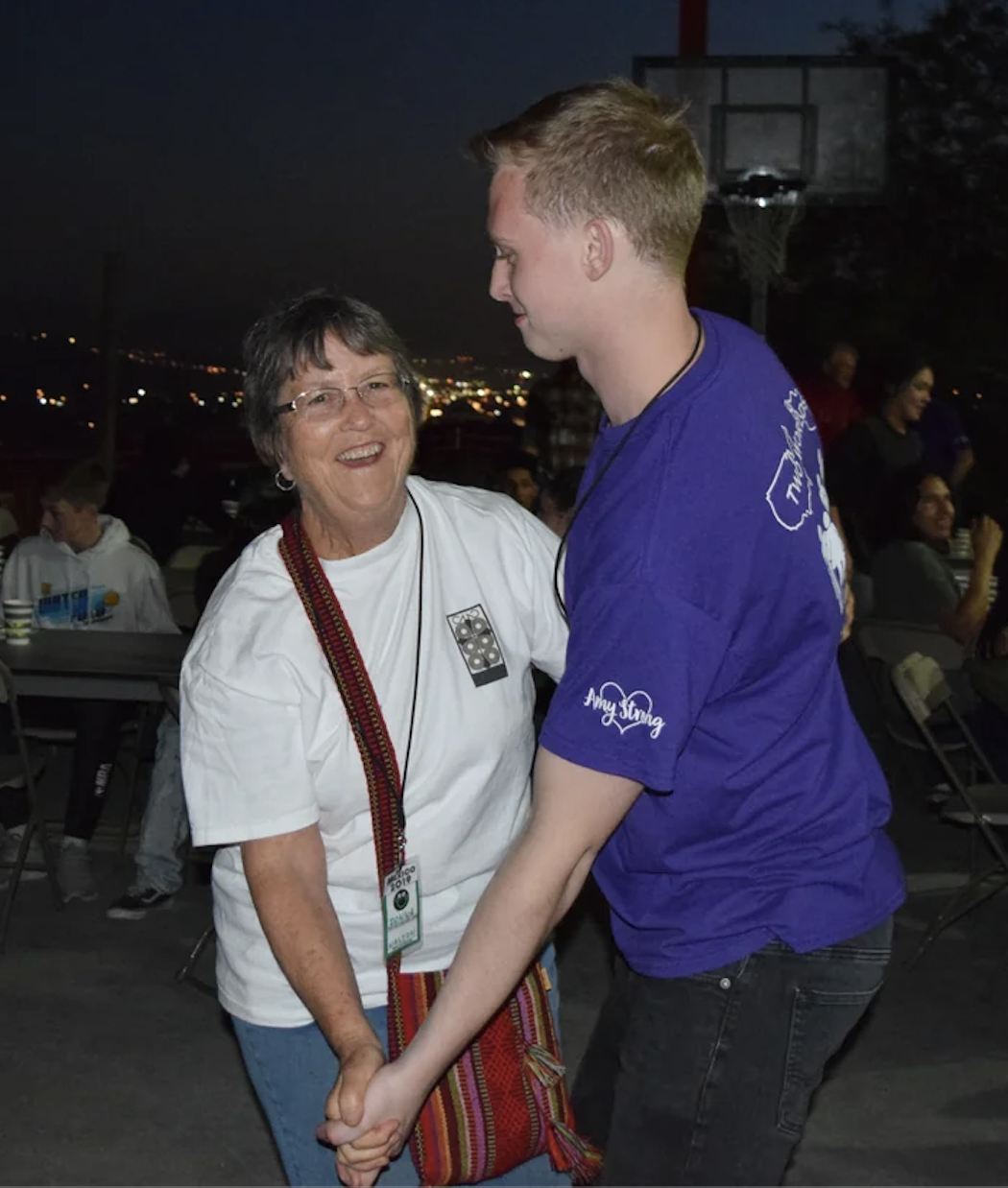 An elderly woman and a young man are dancing and smiling at each other at night, outdoors with a basketball hoop and strings of lights in the background.