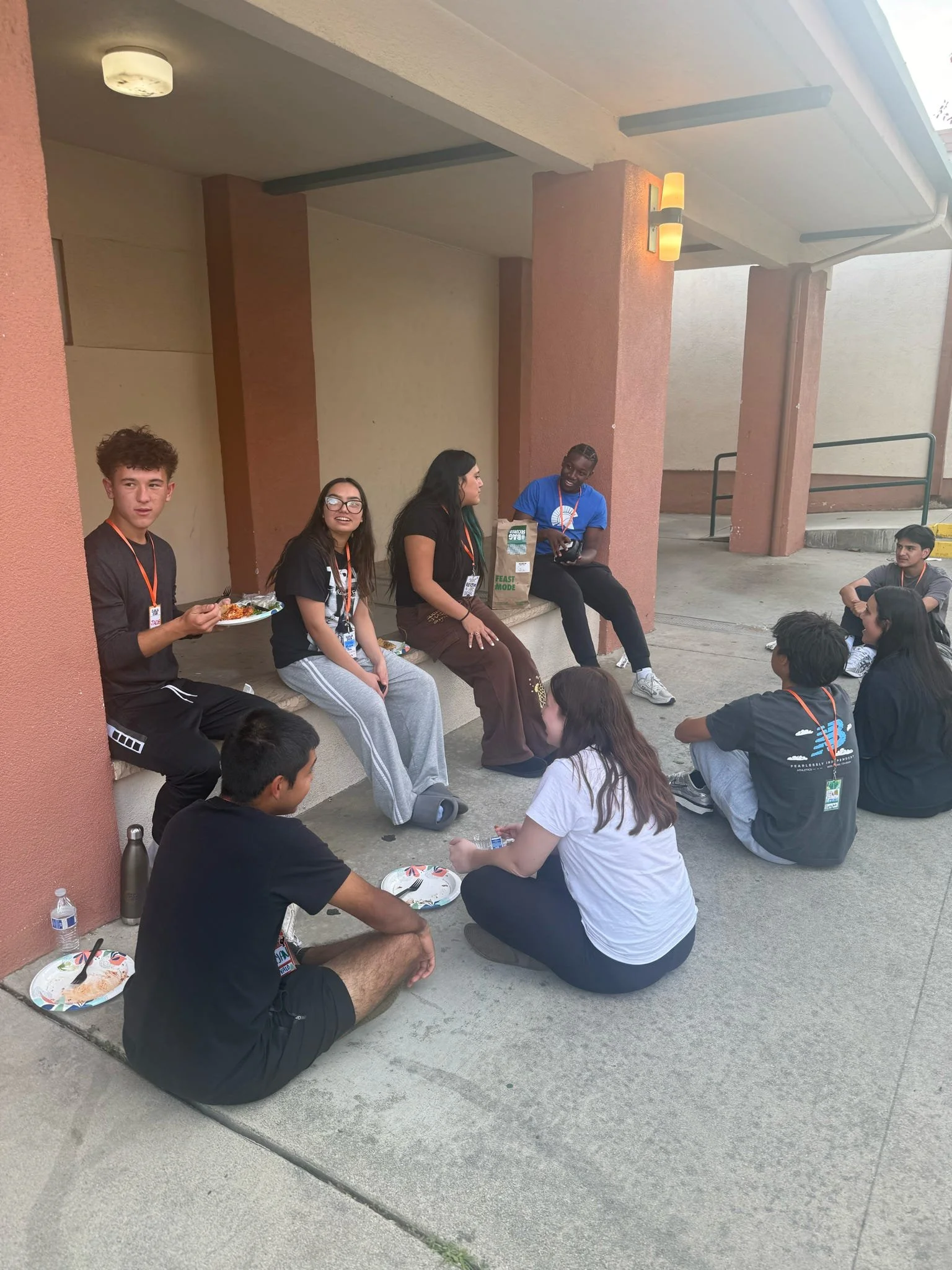 People sitting and chatting outside a building, some with plates of food and water bottles, in a casual social setting.