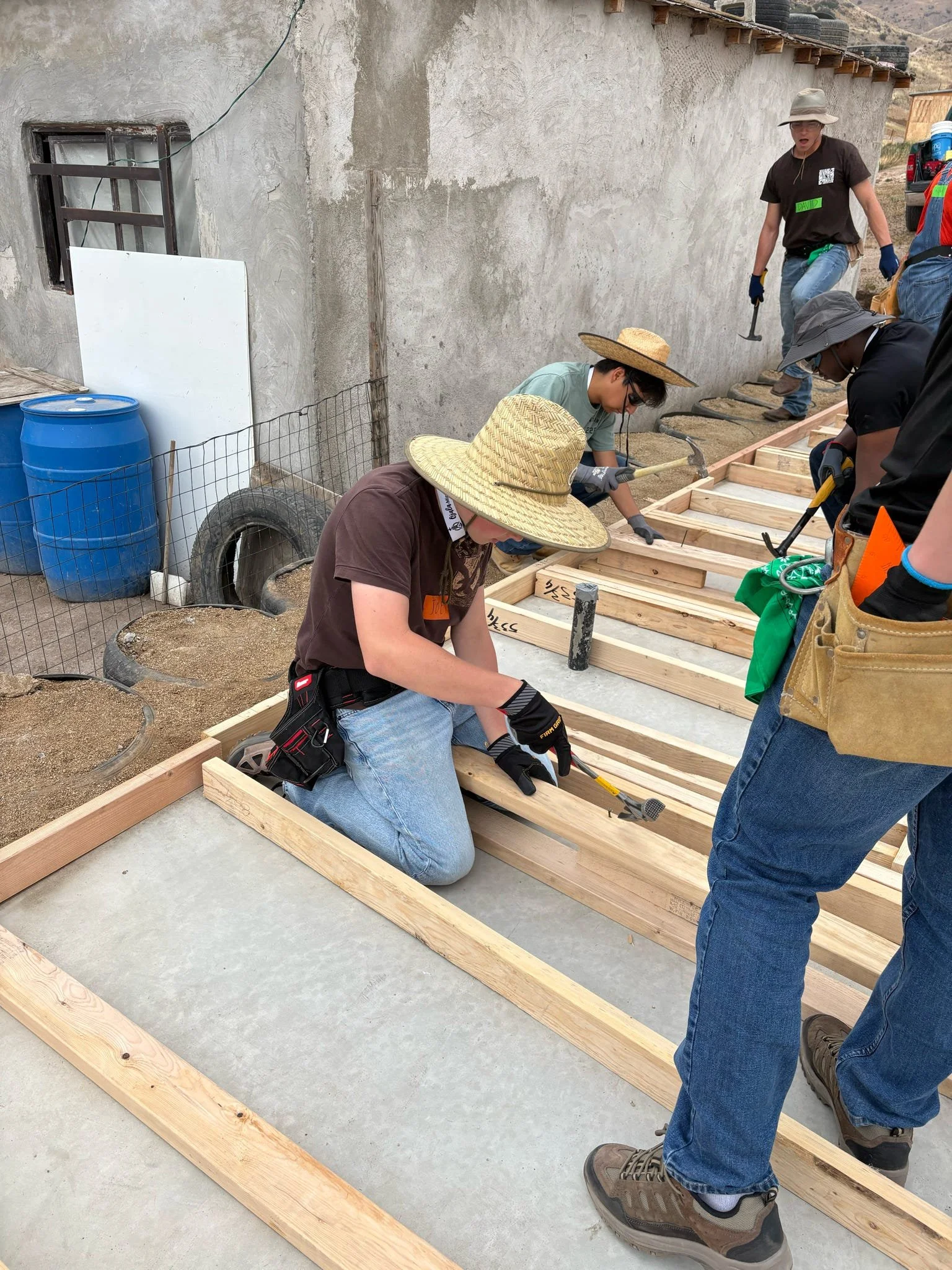 Group of people working on building a wooden frame on a concrete surface outside a weathered building, some wearing hats and safety gear.