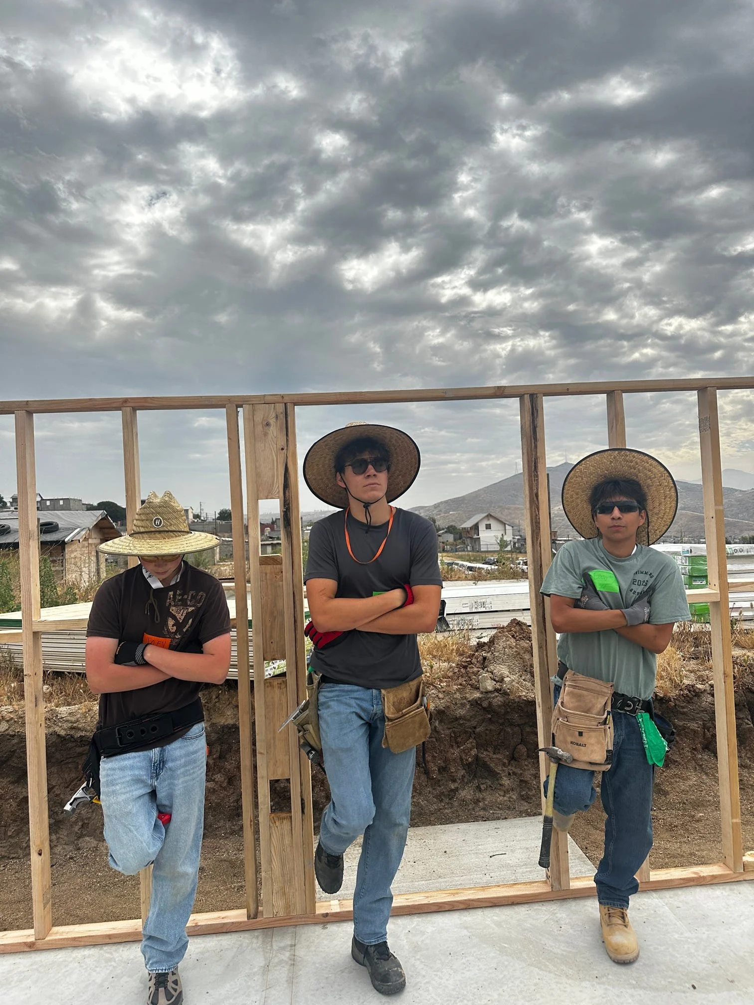 Three teens standing in front of a wooden frame at a building site under cloudy sky. They are wearing protective gear, hats, and sunglasses.