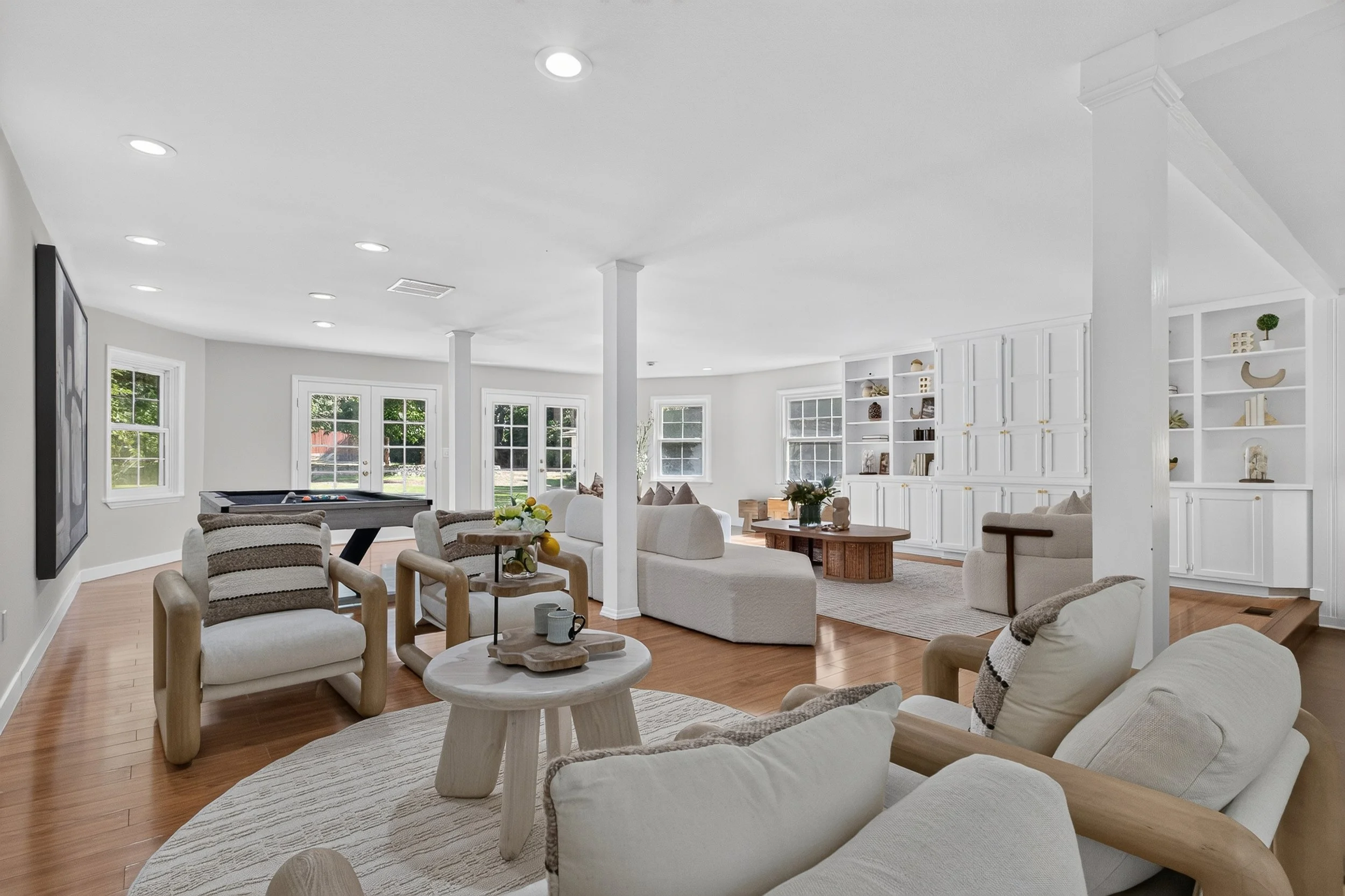 Bright and spacious living room with white walls, multiple windows, wooden flooring, white couches, a pool table, and decorative items on white built-in shelves.