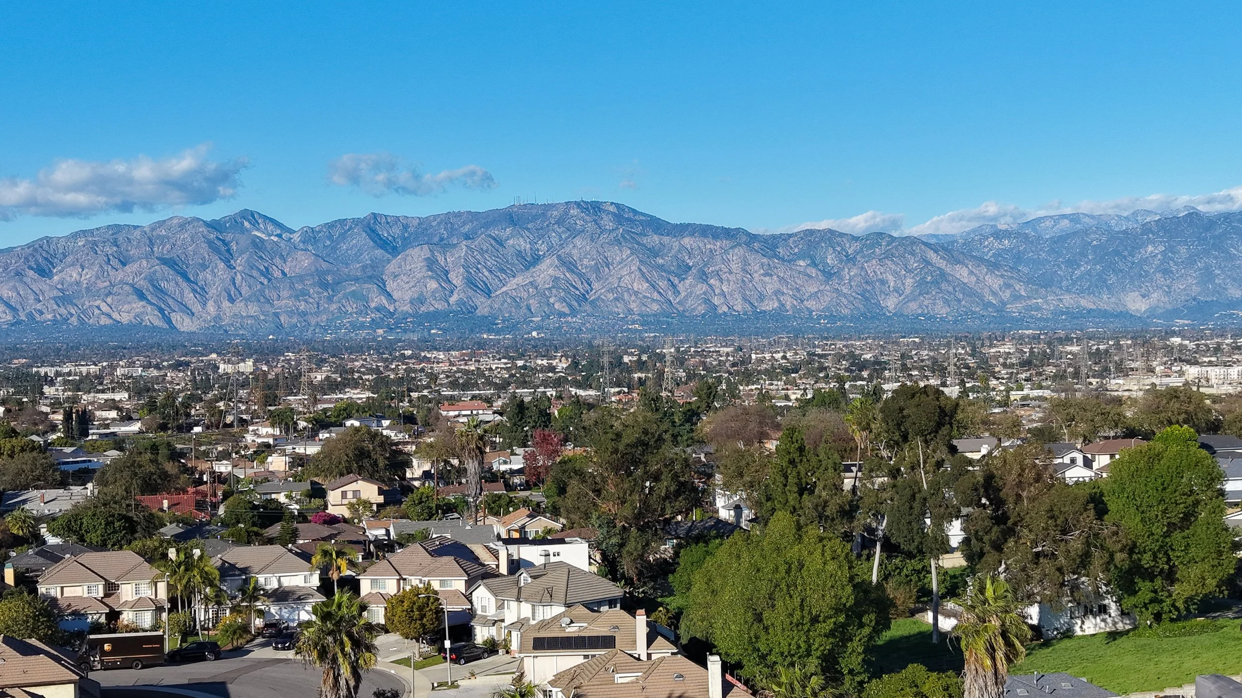 Cityscape with residential neighborhood in foreground, trees, and a mountain range in the background under a clear blue sky with a few clouds.