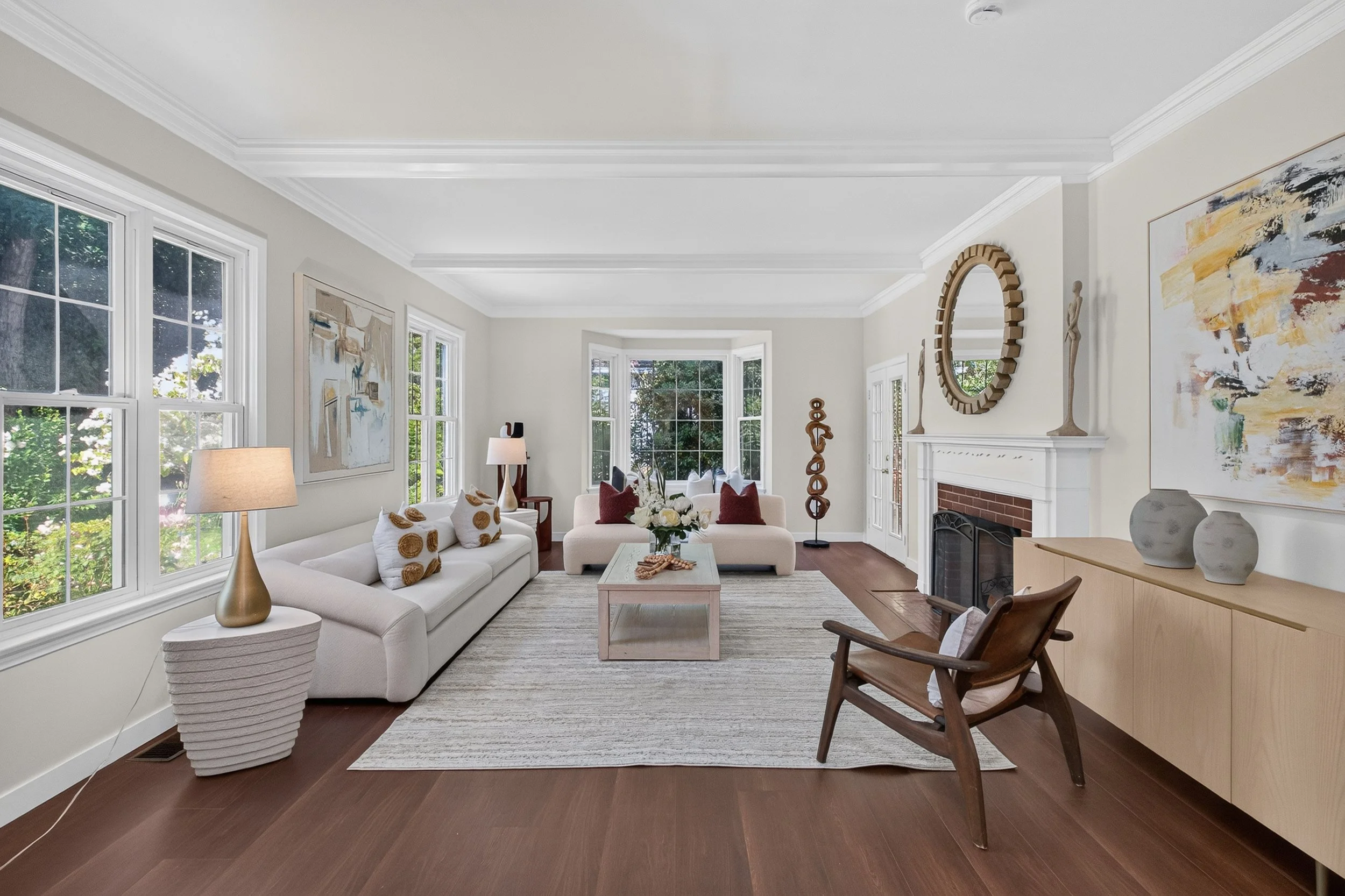 Living room with large windows, white sofa with patterned pillows, a wooden armchair, abstract artwork on walls, a fireplace, and decorative items including a mirror and vases.