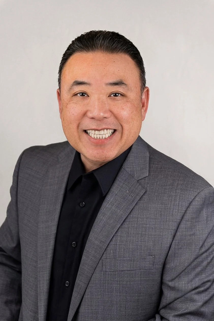 Headshot of a smiling Asian man in a gray suit and black shirt against a plain light background.