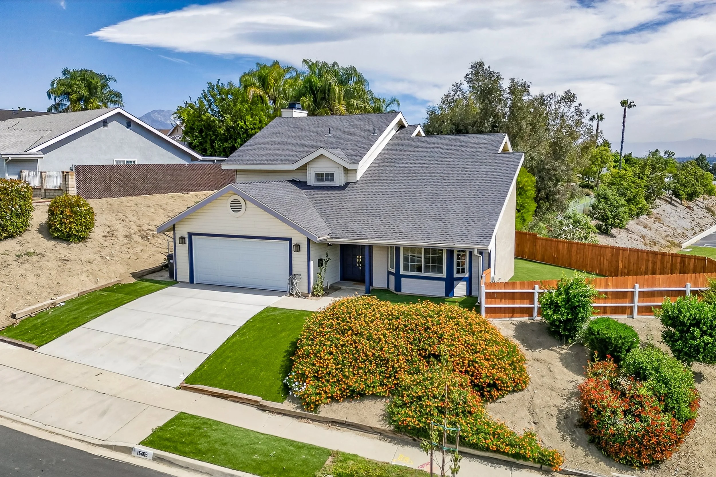 A suburban house with a gray roof, white walls, and a blue garage door, surrounded by lush green grass, colorful bushes, and trees, with a clear sky and some clouds overhead.