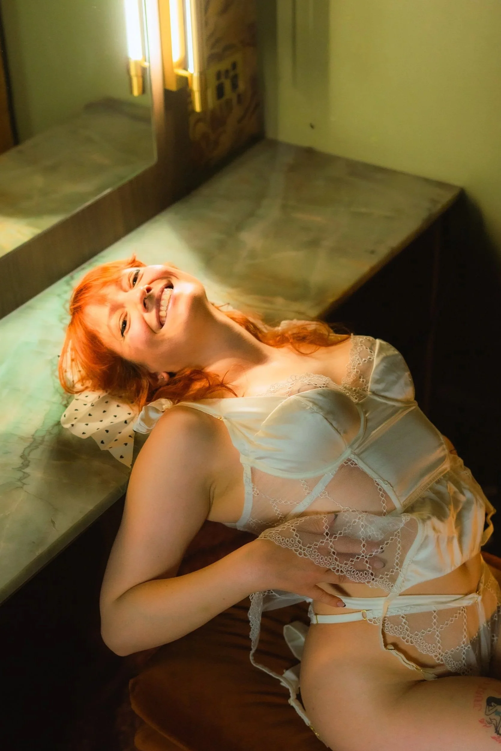A woman with red hair lying back, smiling, wearing white lace lingerie.