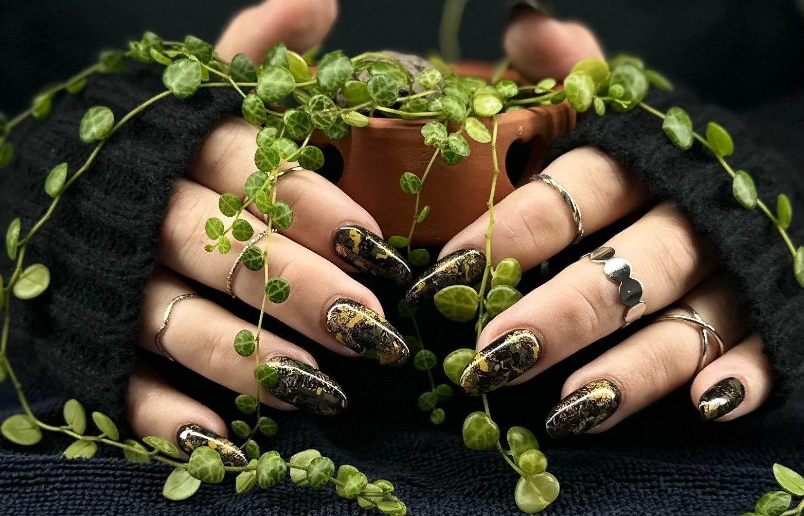 Close-up of bespoke almond-shaped nails featuring black and gold textured nail art, surrounded by trailing green foliage.