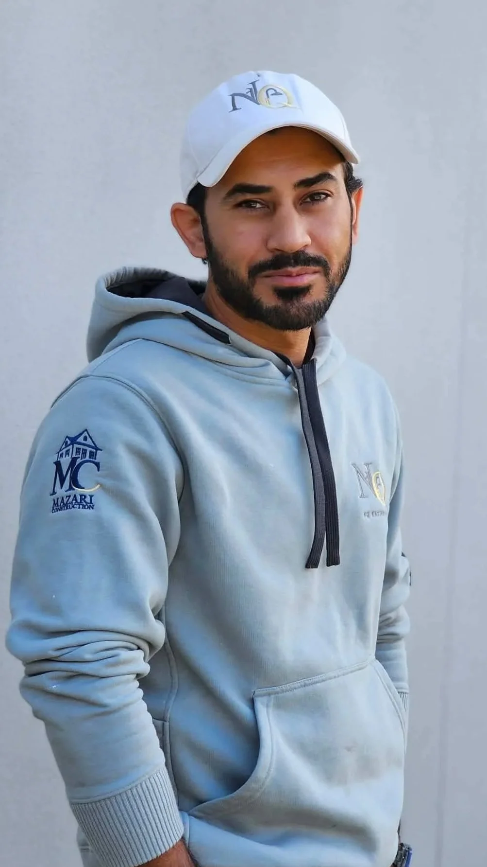 A man with a beard wearing a white baseball cap and a light gray hoodie with logos on the chest and sleeve, standing against a plain white background.