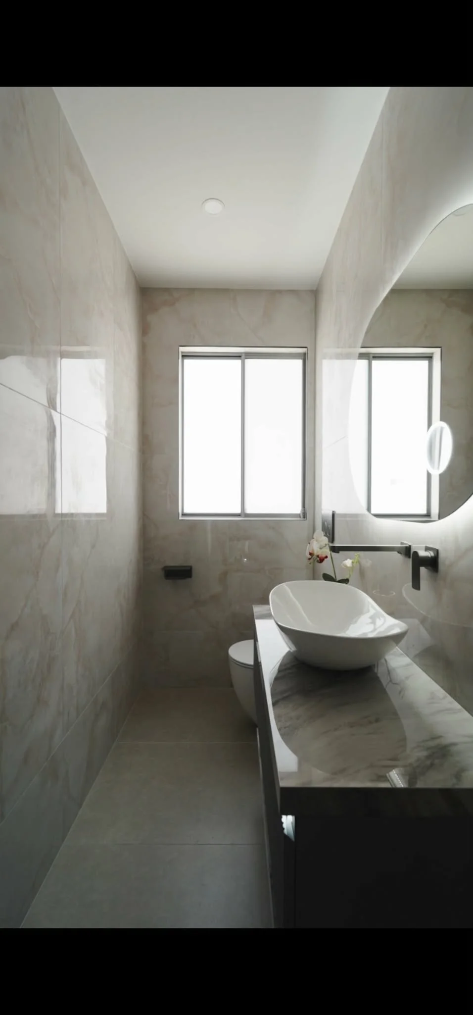 Modern bathroom with marble walls and a vessel sink on a marble countertop, two large windows, a round mirror, and minimal decor.