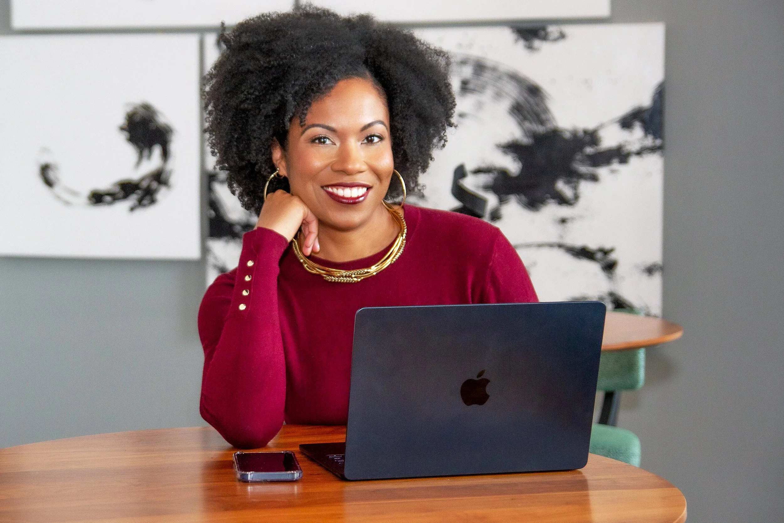 A woman with curly hair wearing a red top and gold jewelry sitting at a wooden table with a laptop and phone, smiling at the camera in a modern room with abstract black and white art in the background.