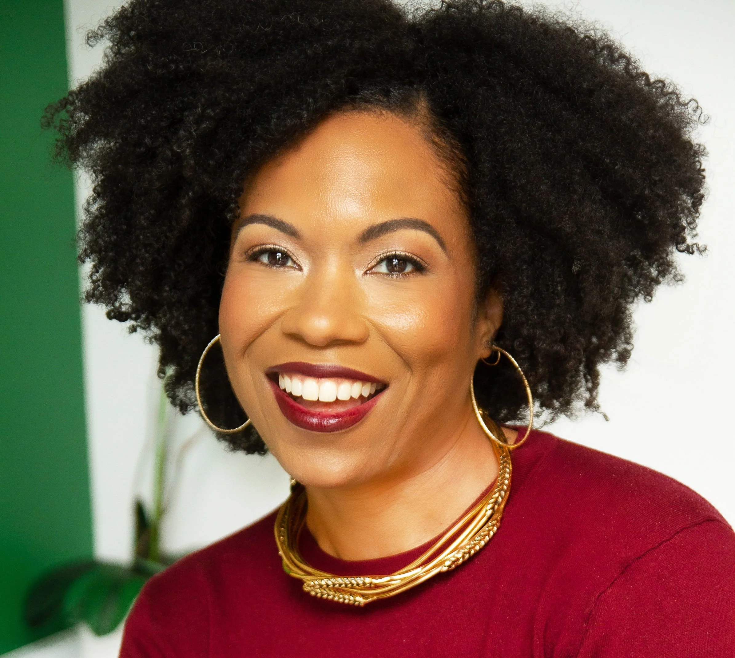 Close-up of a smiling woman with dark curly hair, wearing gold hoop earrings and layered gold necklaces, dressed in a red top, with a green and white background.