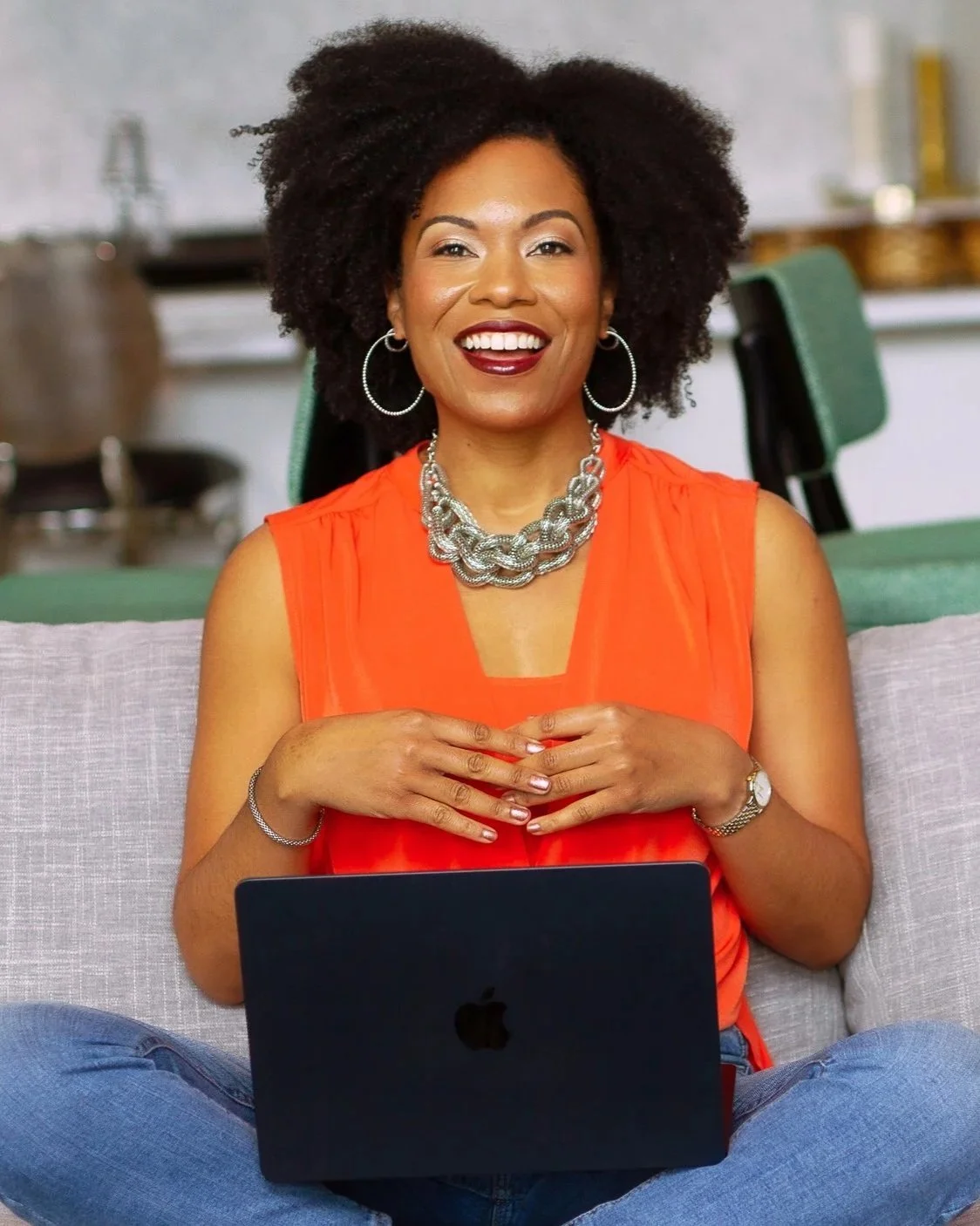 A young woman with curly hair smiling, wearing an orange sleeveless top and jewelry, sitting on a couch with a laptop on her lap, in a casual indoor setting.