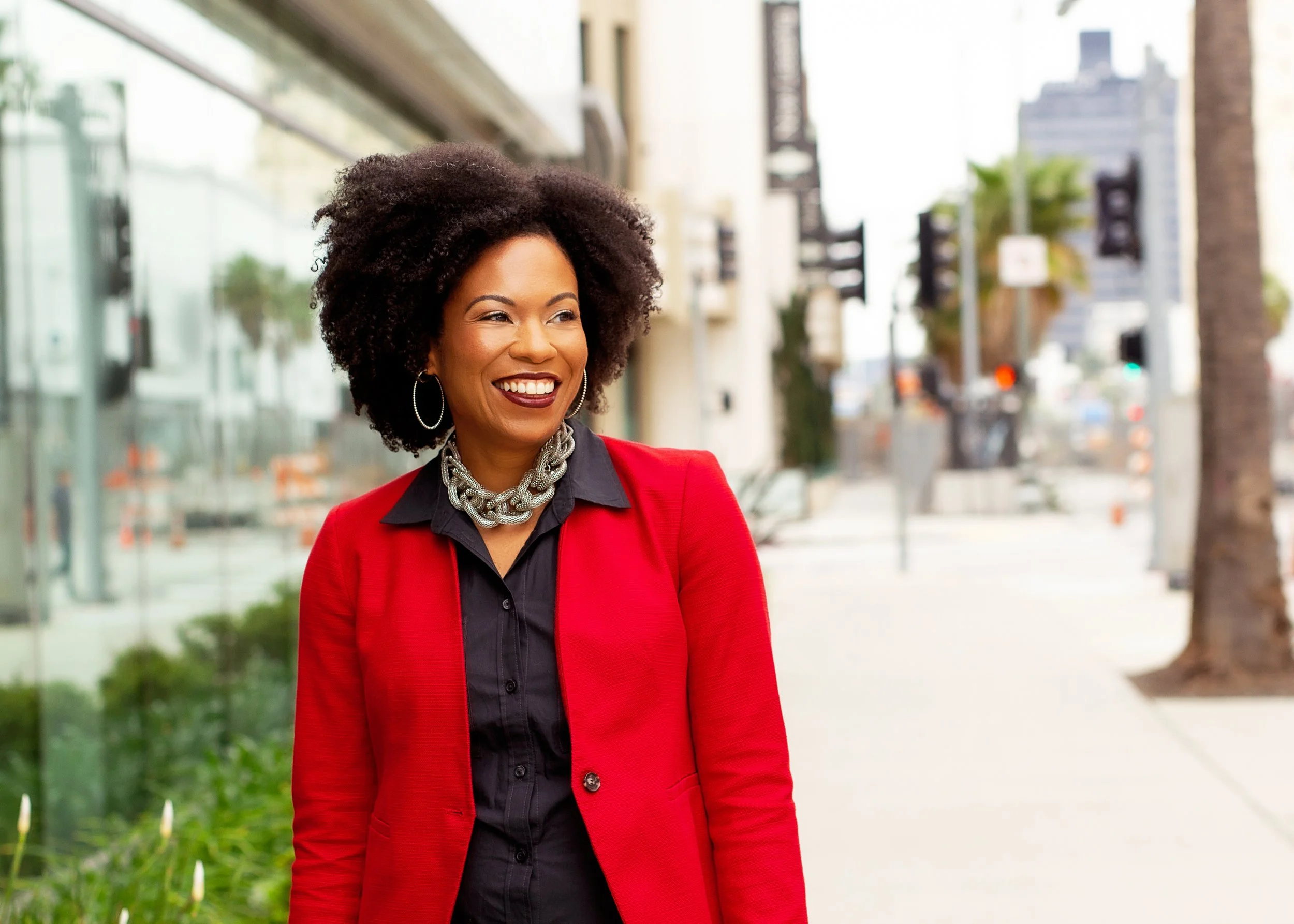 A woman with dark curly hair, wearing a red blazer and a black shirt, smiling while walking on a city sidewalk with buildings and palm trees in the background.