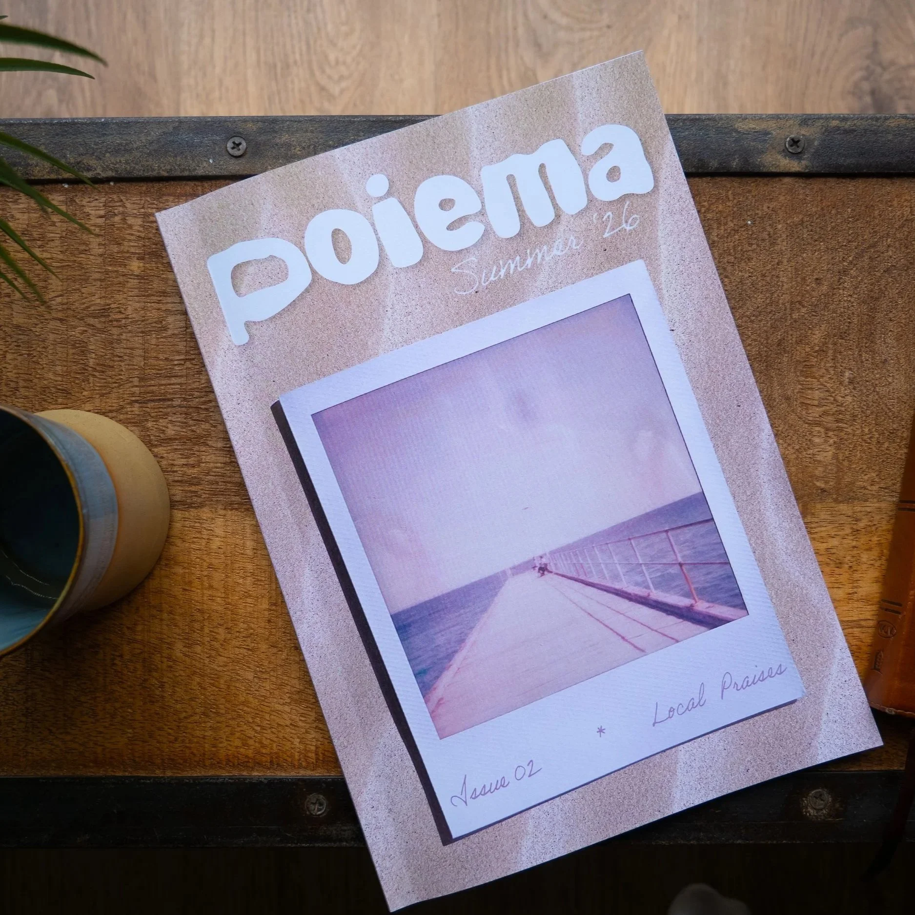 Close-up of a printed flyer with the word 'Poiesis' in large white letters on a pink background, titled 'Summer 26,' with a faint image of a bridge or pier over water and handwritten notes at the bottom, resting on a wooden surface next to a yellow and black mug.