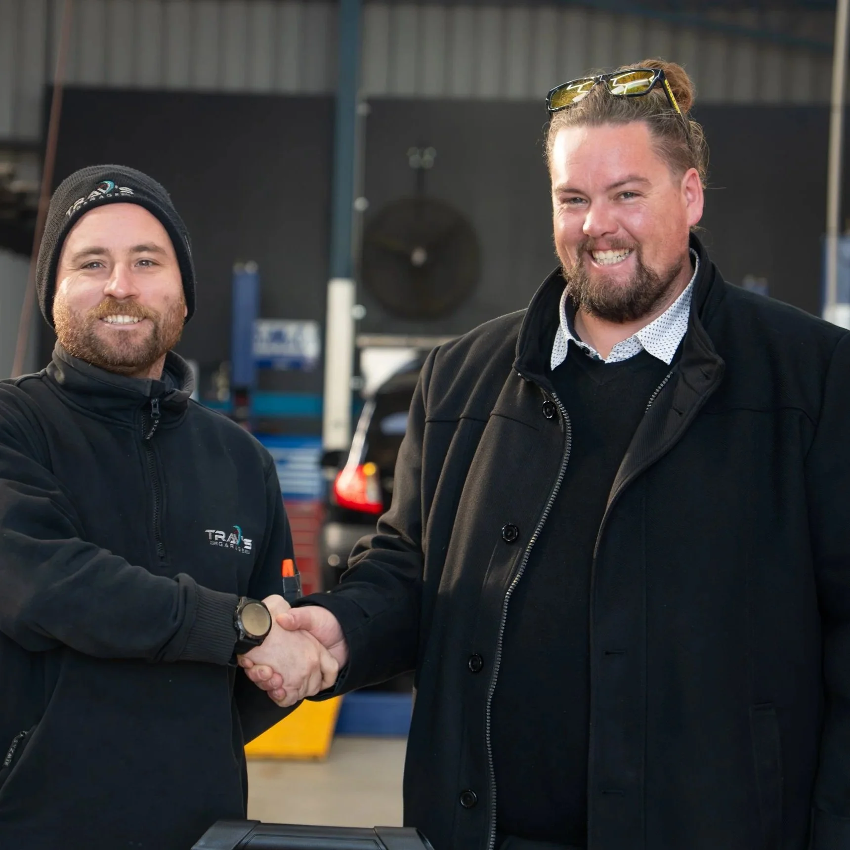 Two men shaking hands inside a garage, one wearing a beanie and the other with glasses on his head, both smiling.