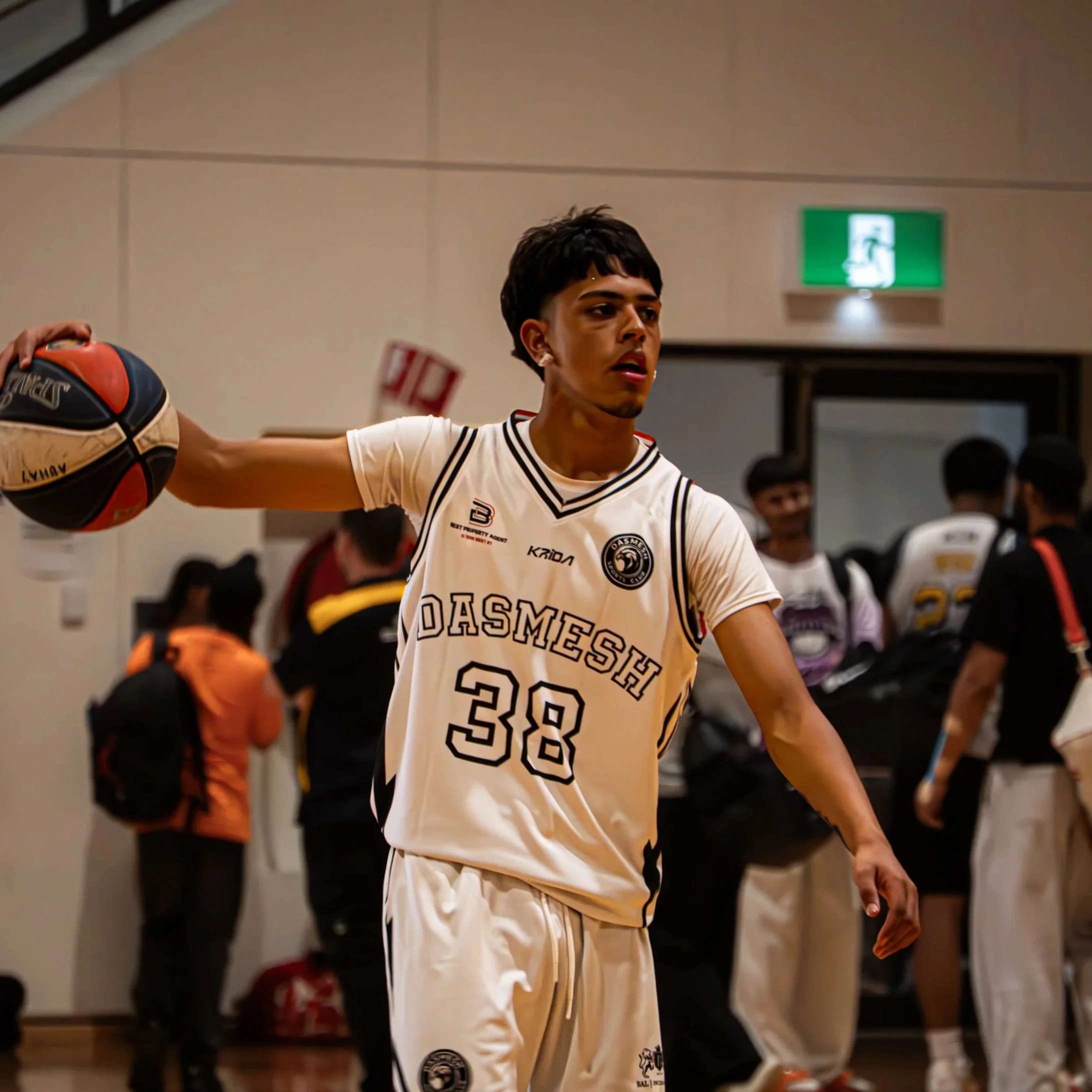 Young male basketball player in a white DASMEIS uniform holding a basketball, in a gymnasium with others in the background.