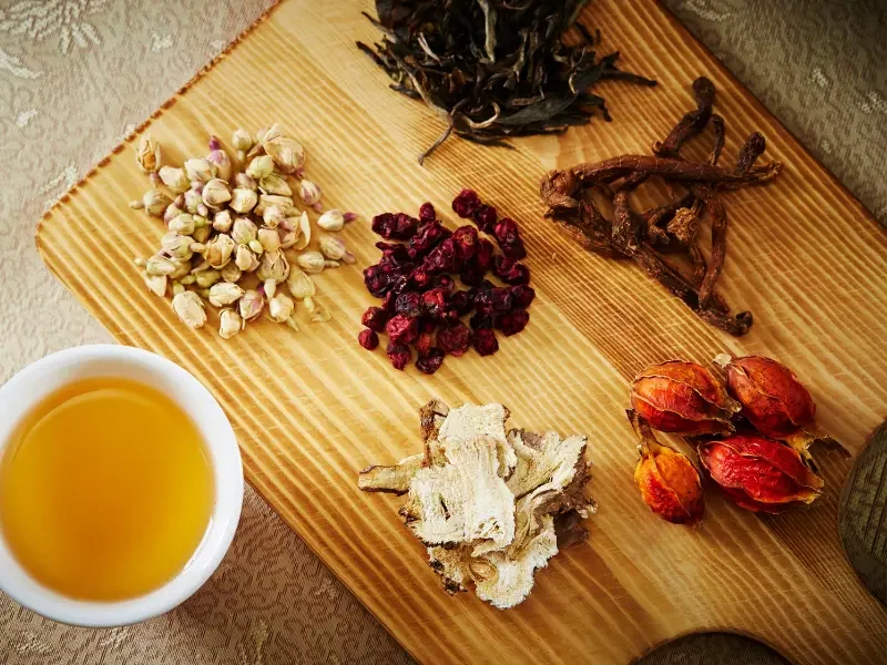 Herbal medicine ingredients including dried roots, berries, and flowers arranged on a wooden board with tea