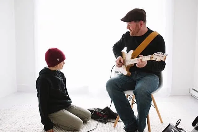 A man playing an electric guitar while a young boy listens, both sitting and facing each other in a bright room with white curtains.