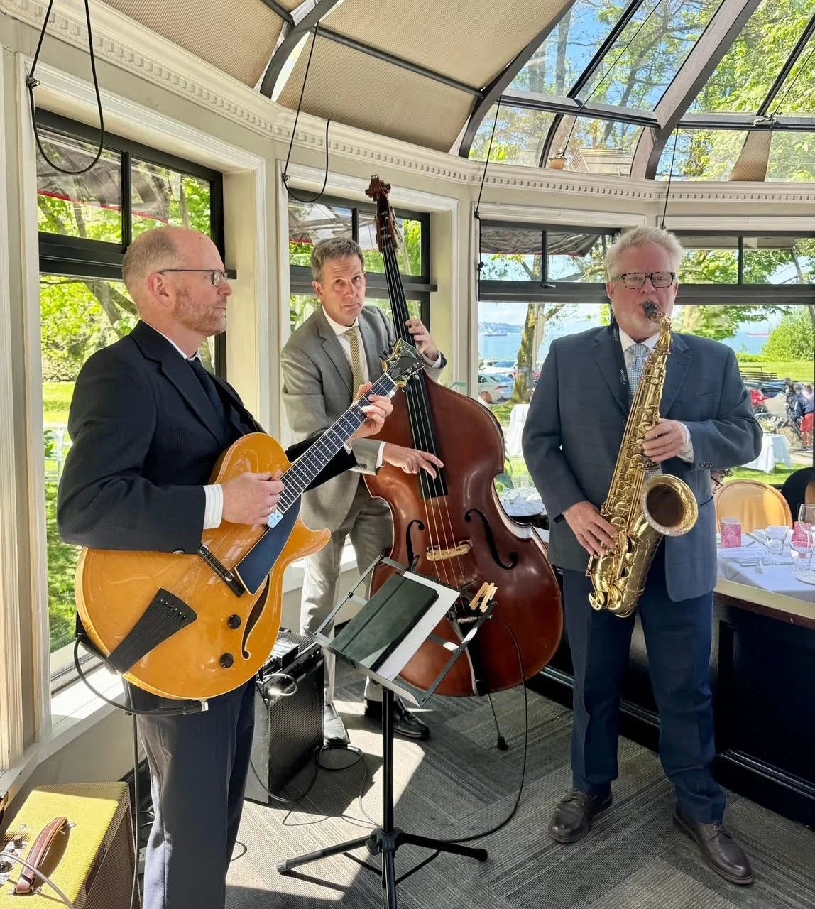 Jazz band performing indoors with three musicians playing guitar, double bass, and saxophone, in a bright room with large windows and a glass ceiling showing trees outside.