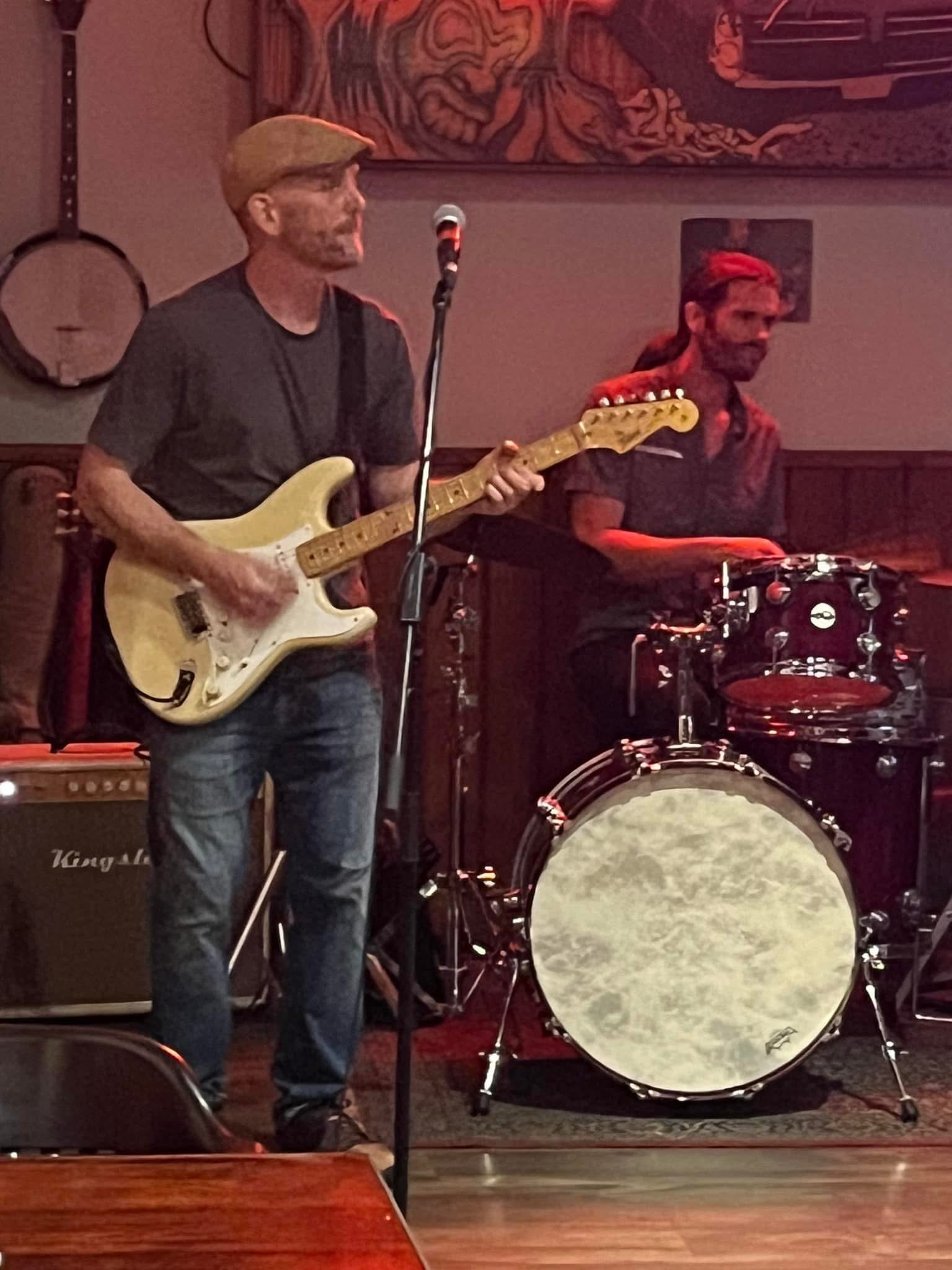 Two musicians perform in a dimly lit venue. One is playing an electric guitar and the other is on drums. The guitarist has a beard, short hair, and is wearing a black t-shirt and a flat cap. The drummer has a beard, long hair tied back with a bandana, and is wearing a short-sleeved shirt.