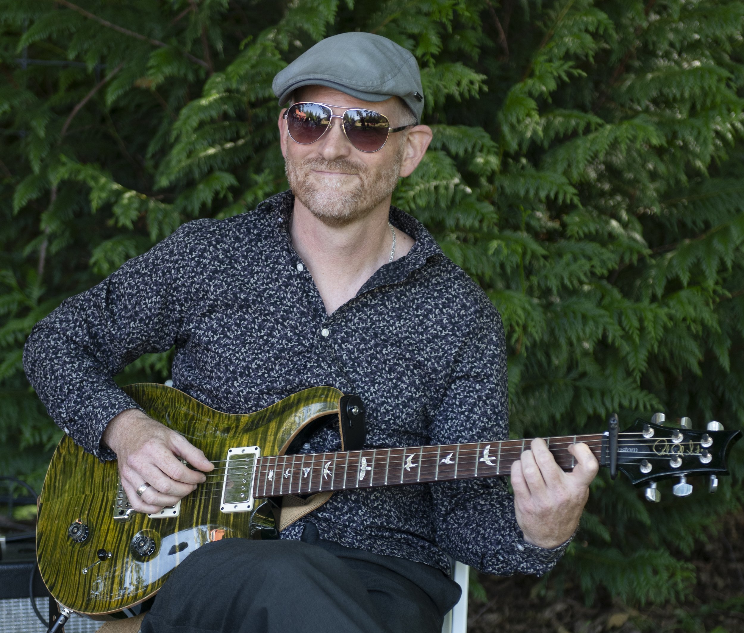 A man wearing sunglasses, a gray cap, and a dark patterned shirt is playing an electric guitar outdoors in front of dense green foliage.