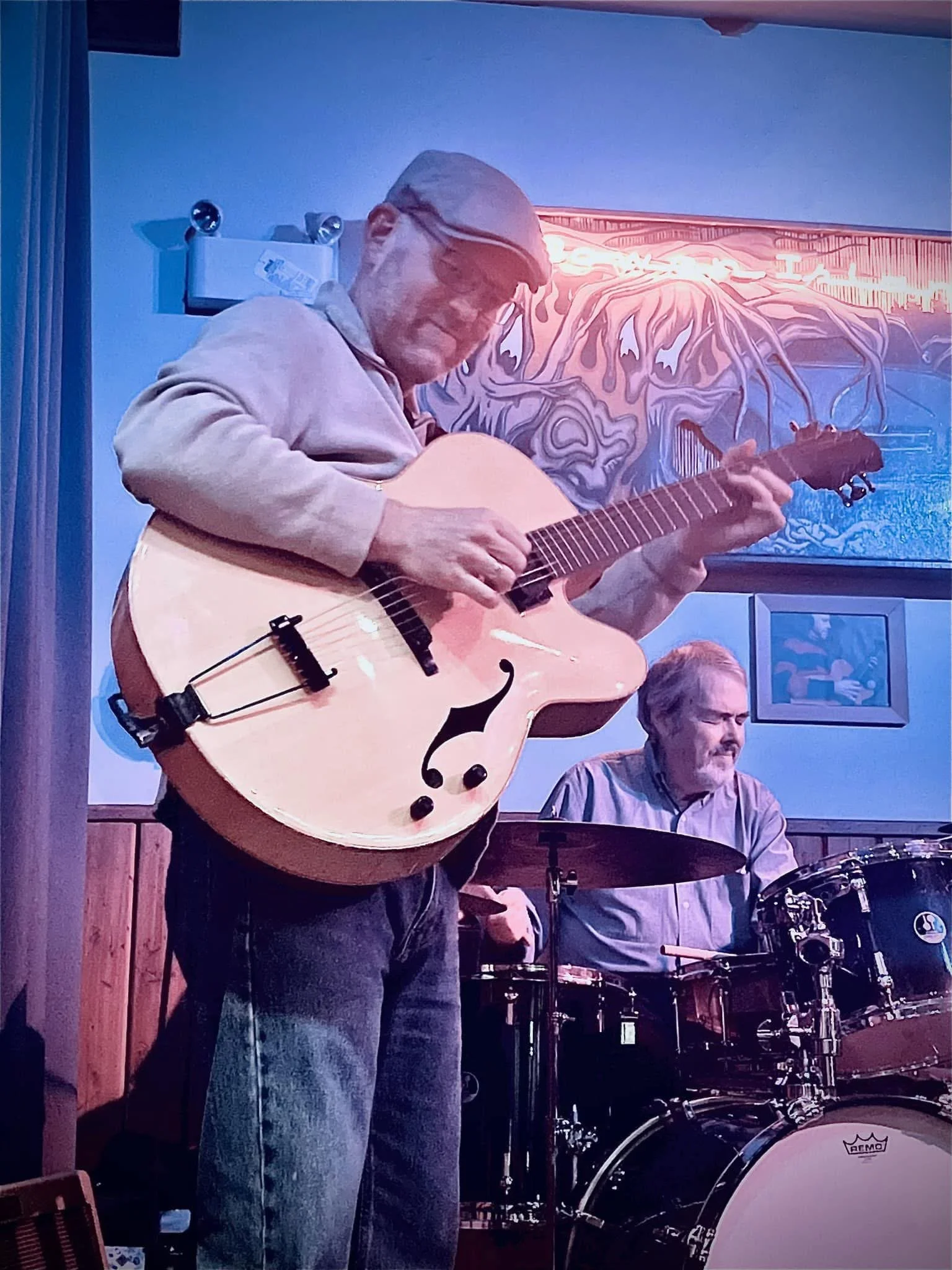 A man playing an acoustic guitar with a musician playing drums in the background, inside a room with artwork on the wall.