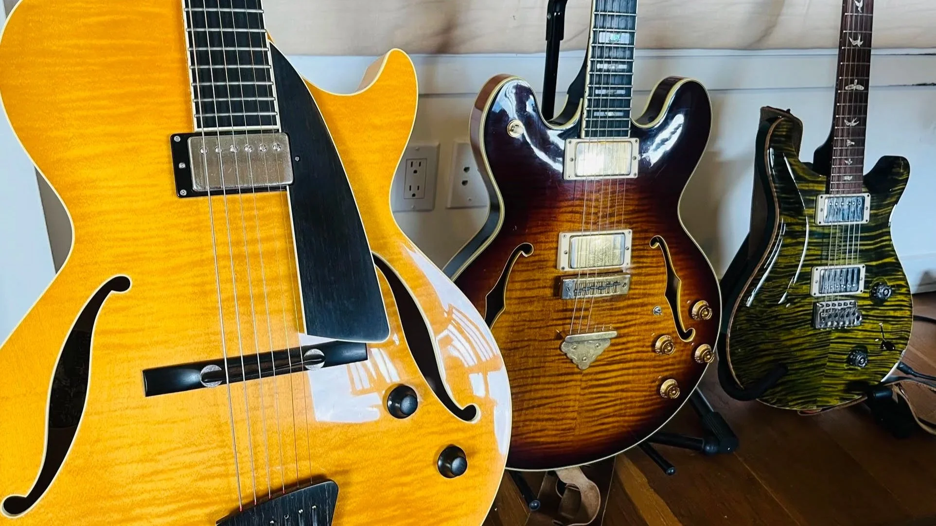 Three electric guitars with different finishes and shapes, leaning against a wall with electrical outlets visible in the background.