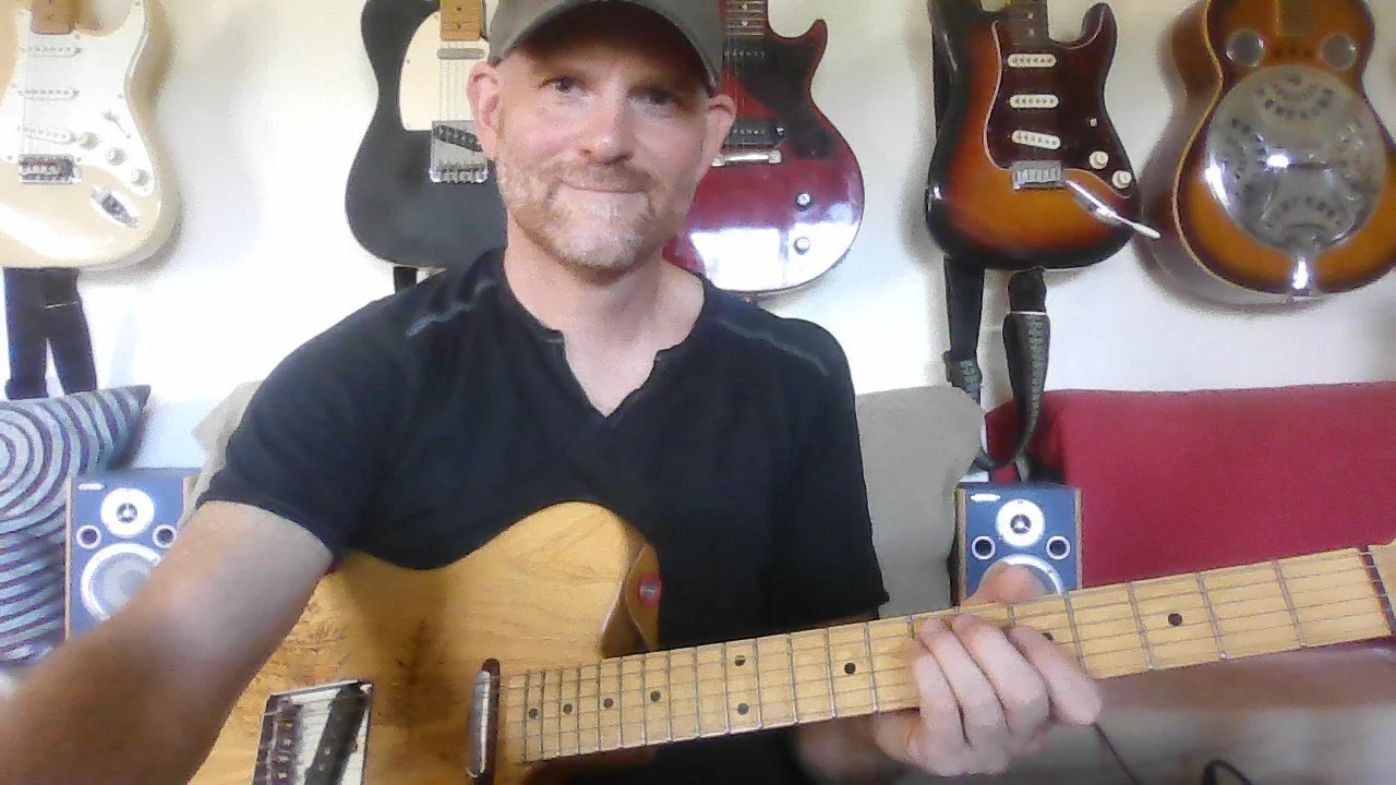 A man wearing a black shirt and a cap holding an electric guitar in a room with several guitars hanging on the wall behind him.