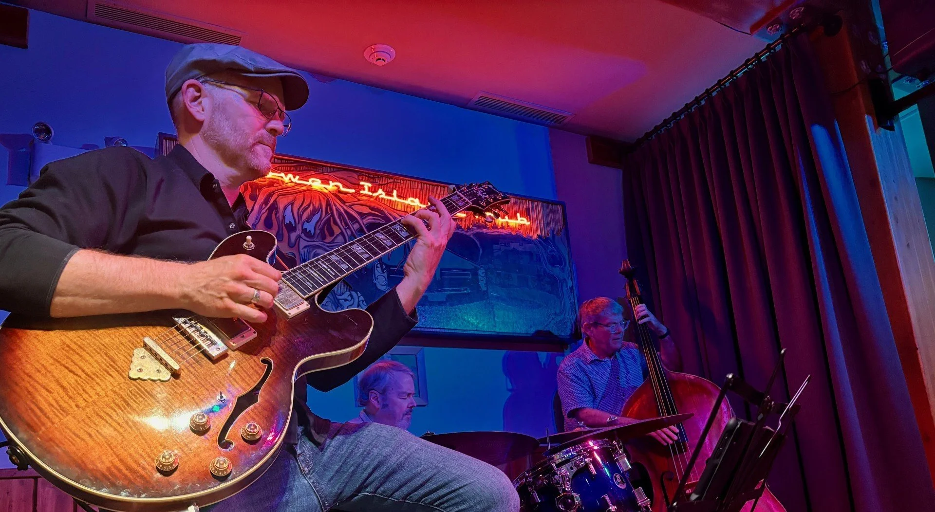 Jazz band performing live, with a guitarist in the foreground and two other musicians playing bass and drums in a dimly lit bar or venue.