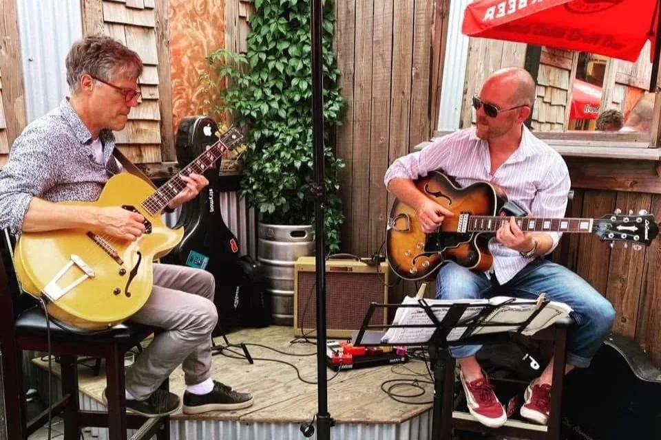 Two men playing guitars and a music stand with sheet music in an outdoor setting with wood fencing and a red umbrella.