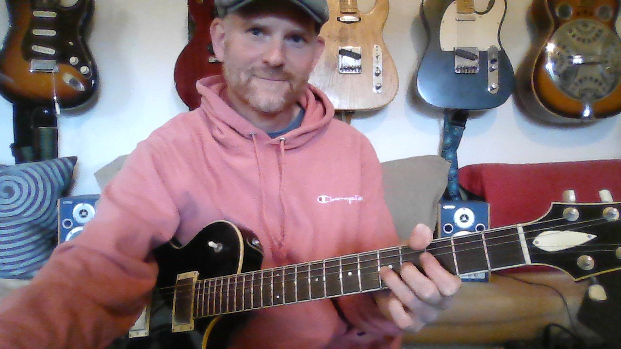 A man wearing a pink hoodie and a cap playing an electric guitar in a music studio with guitars hanging on the wall behind him.