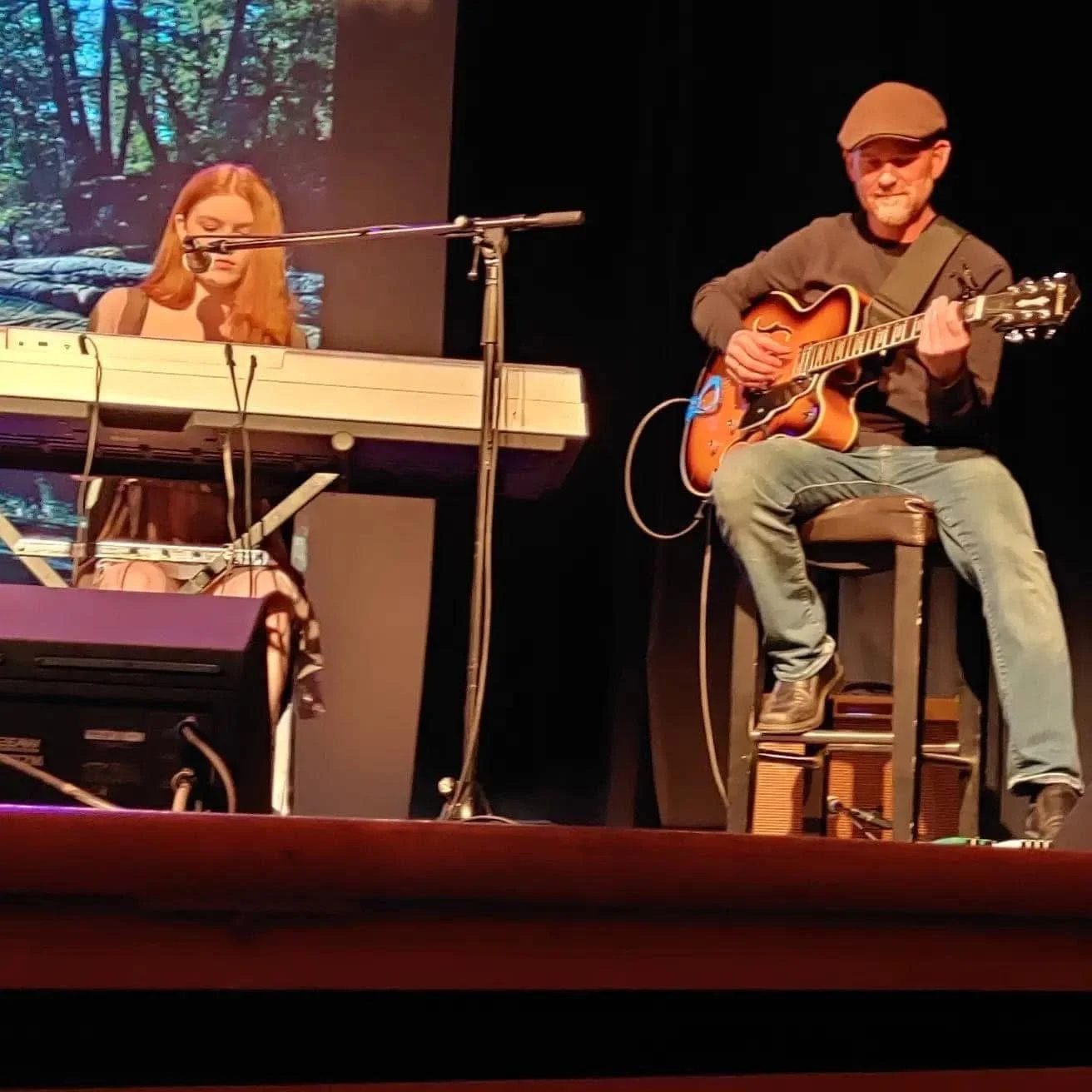 A man playing an electric guitar and a woman playing a keyboard on a stage. The background has a nature scene projected on a screen.