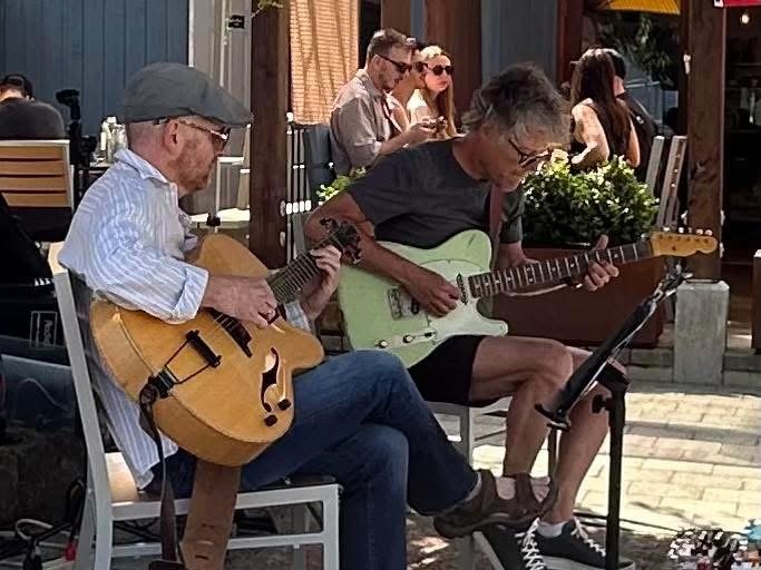 Two men playing acoustic guitars outdoors on a sunny day, with several people sitting at tables in the background.
